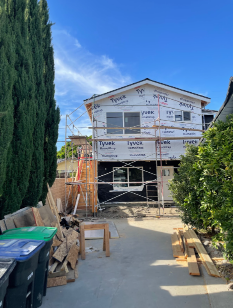 House under construction with scaffolding, construction materials on the ground, and a clear blue sky.