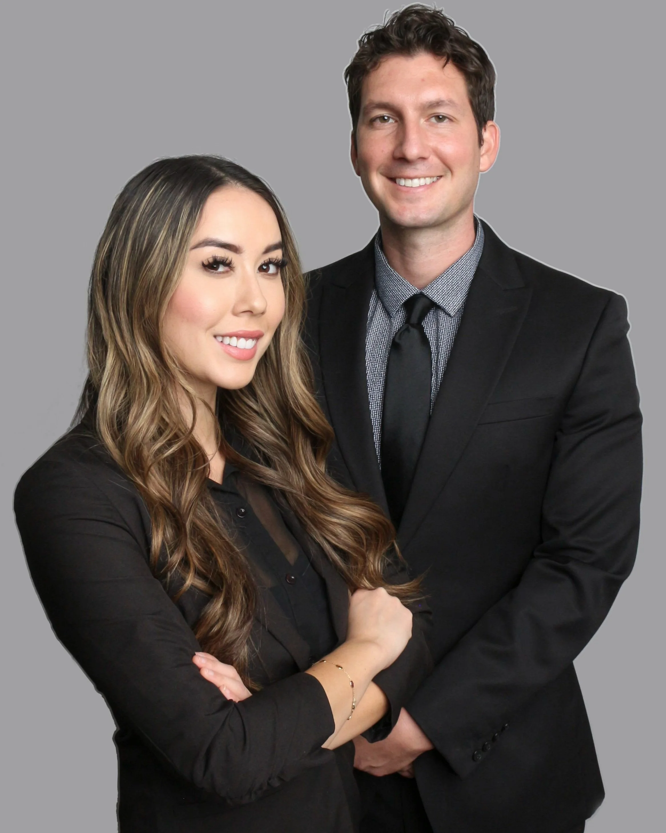 A smiling woman with long wavy brown hair and dark eye makeup, wearing a black top, standing with crossed arms. A smiling man with short dark hair, wearing a black suit, gray dress shirt, and black tie, standing with his arms crossed. Both are posing against a plain gray background.