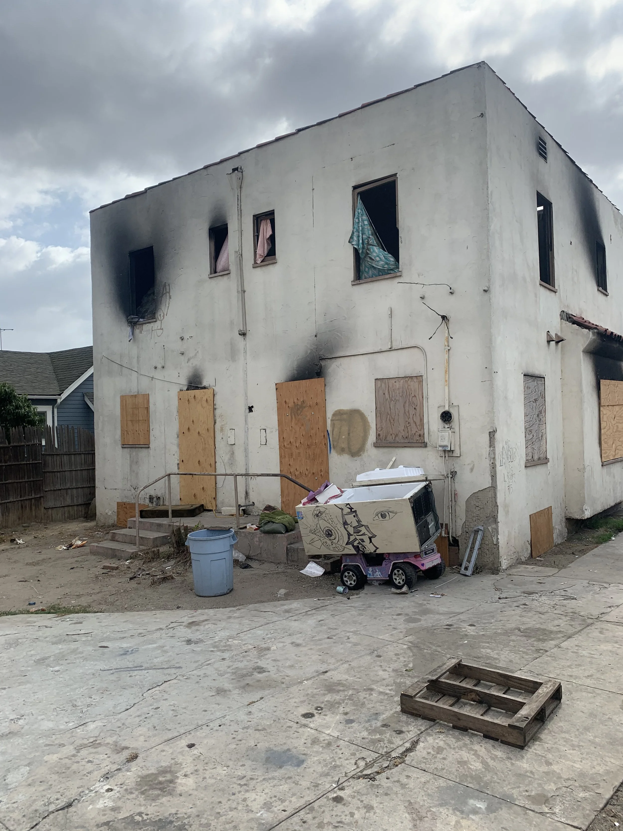 An unfinished, two-story building with boarded-up windows, some open, showing clothing hanging out. The building has a white, weathered exterior with some black soot marks. In front, there is a concrete yard with scattered debris, including a cardboard box with drawings, a wooden pallet, a blue trash can, and random trash. The sky is cloudy.