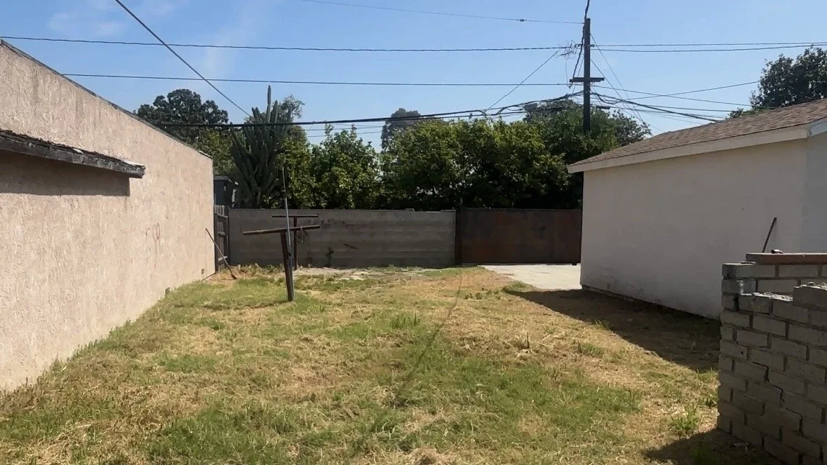 Backyard with grass, a wooden fence, a white house on the right, utility poles and trees in the background under a clear blue sky.
