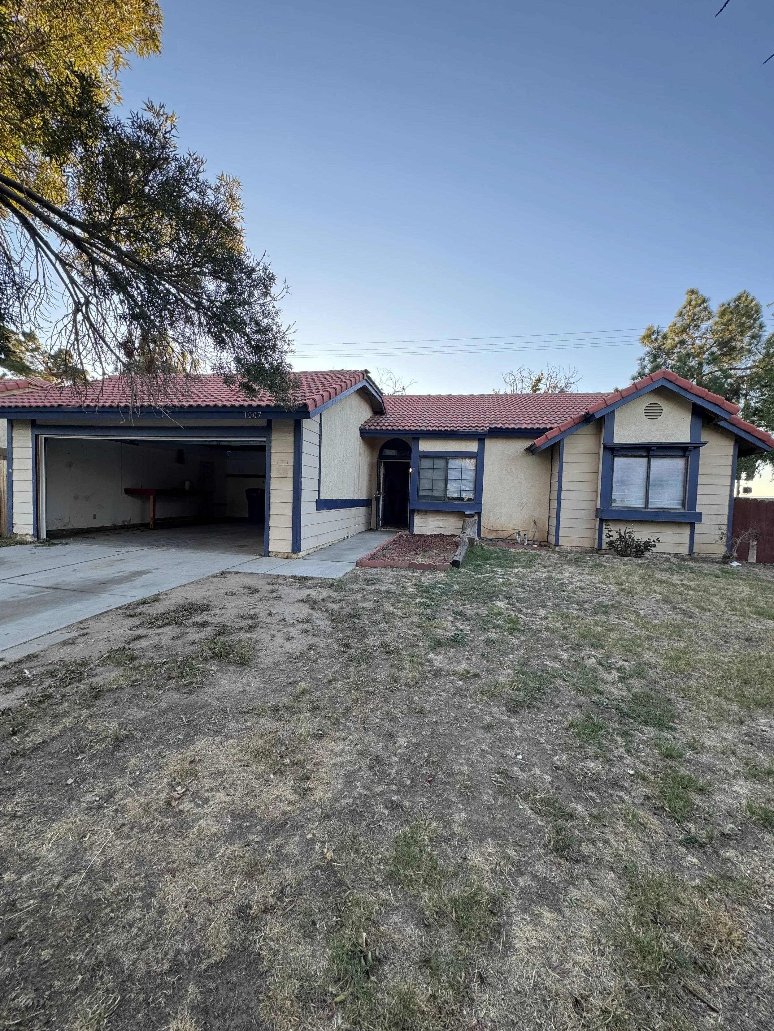 Front yard of a house with a large tree on the left, a driveway leading to an open garage on the left, and a house with a red-tile roof and blue trim. The yard has patches of grass and dirt.