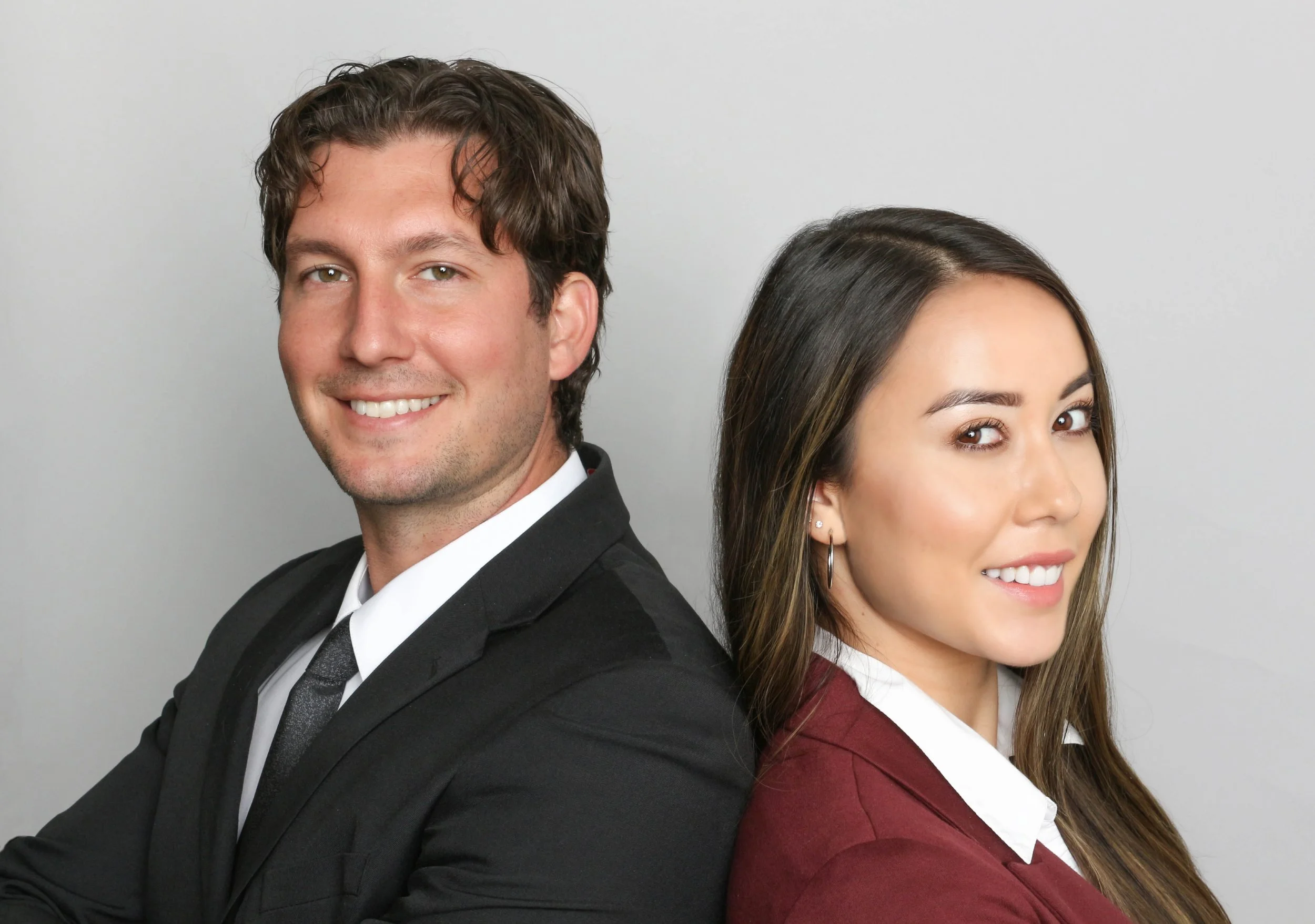 Portrait of a smiling man in a tuxedo and a woman in a burgundy jacket and white shirt, both facing sideways against a plain background.