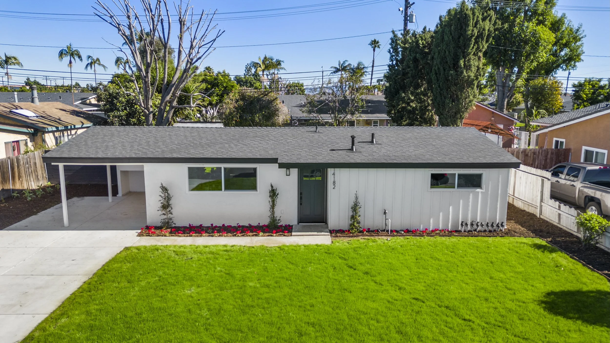 A modern single-story house with a black roof, white exterior walls, and a front yard with green grass and flower beds. There are windows, a front door, a driveway with a car, and trees in the background.