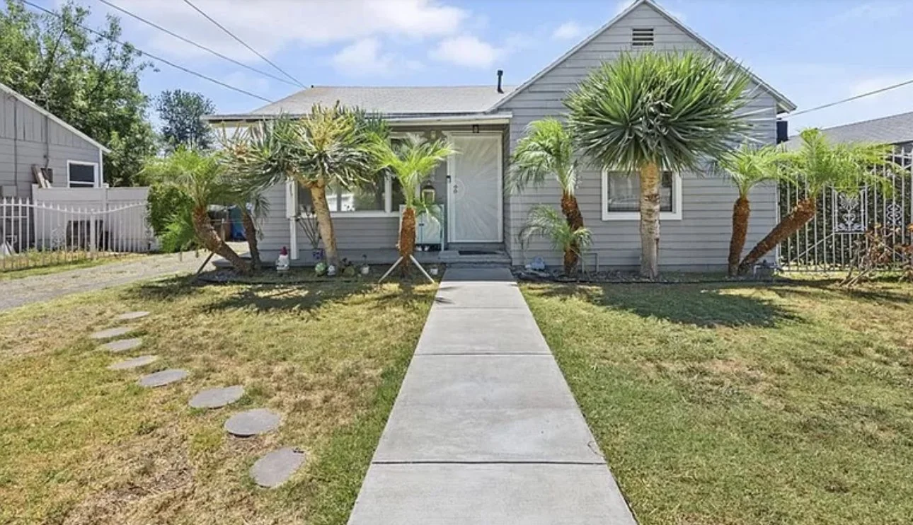 Front yard with a concrete walkway leading to a house entrance, surrounded by palm trees and grass.