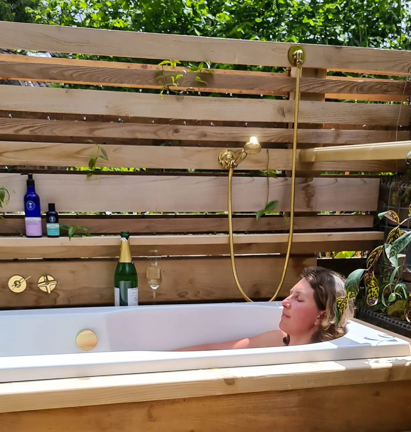 Women relaxing in outdoor bath surrounded by wooden panelling.  A glass of bubbly is by the side of the bath and plants grow in around the bath