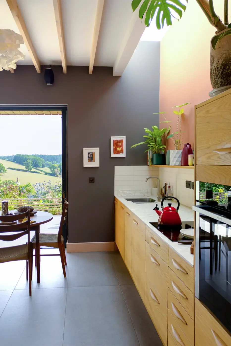 Kitchen with contemporary oak cabinets, a red kettle on the induction hob and a light worktop with inset sink. The shelf above has books, plants and ceramics. All lit from skylight above