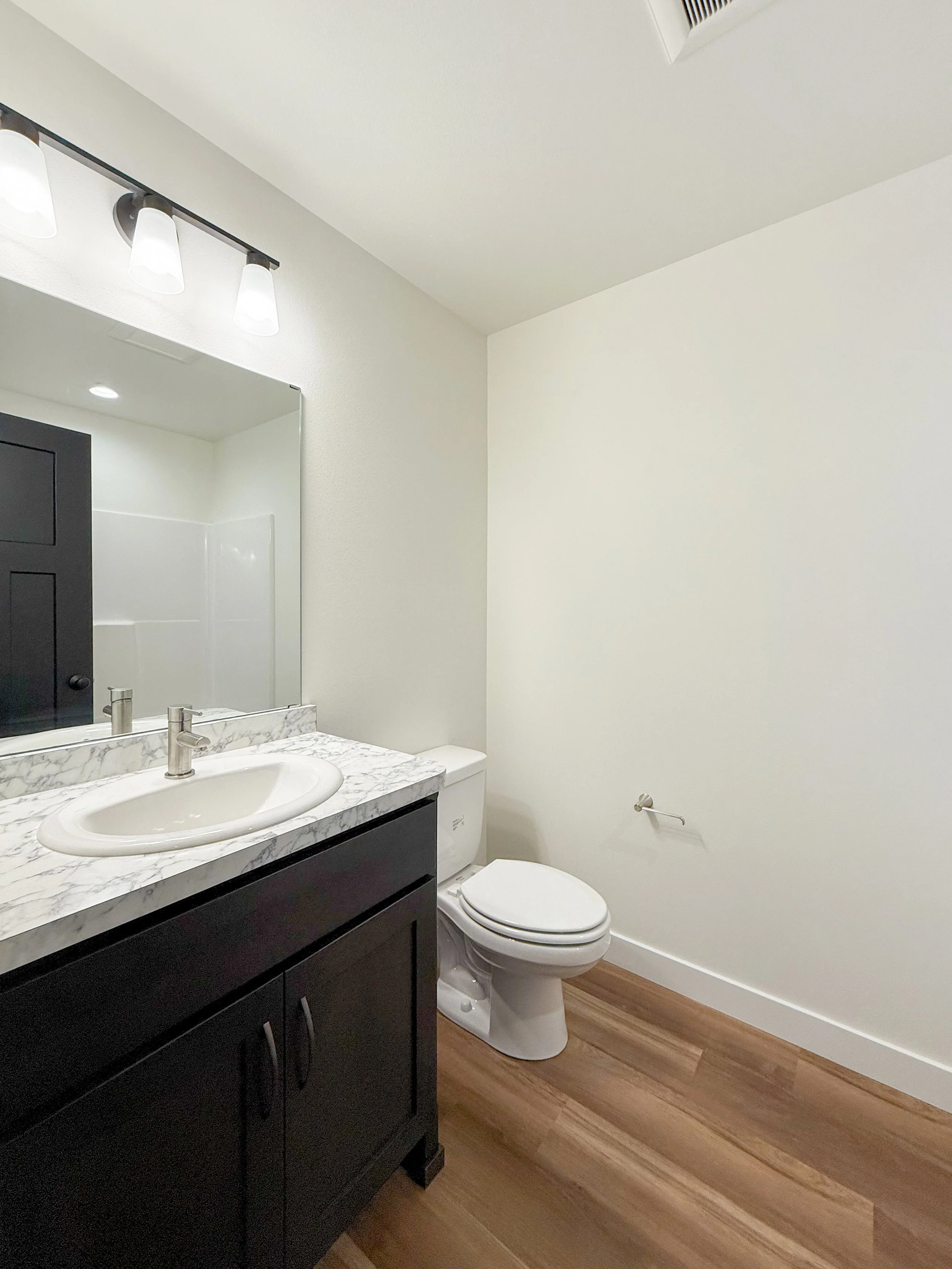 Bathroom with a black vanity, marble countertop, white sink, wall-mounted faucet, large mirror, white toilet, wood flooring, white walls, ceiling light, and vent