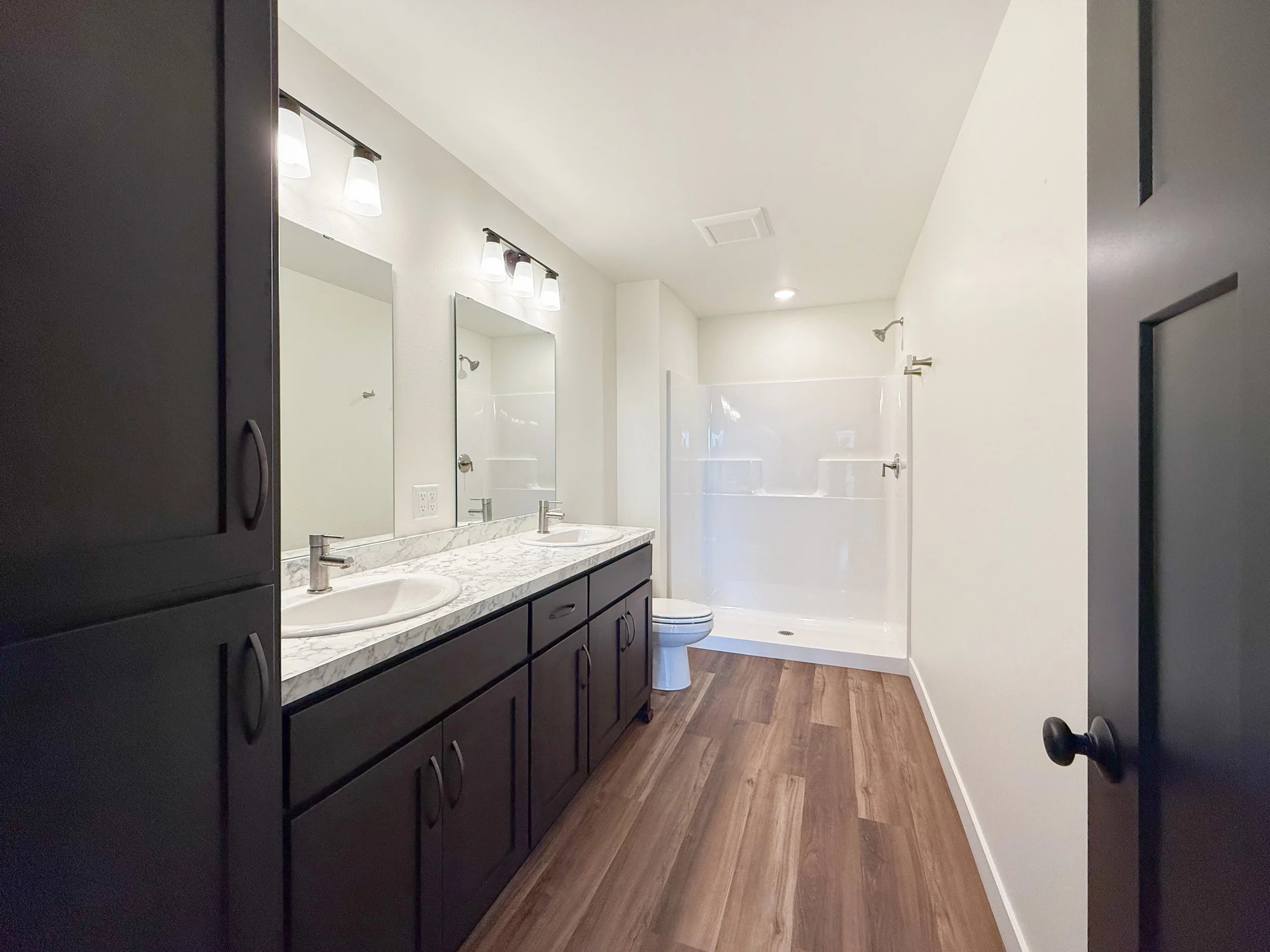 Bathroom with dual sinks, black cabinetry, marble countertop, large mirror, light fixtures, bathtub with glass shower door, and wood-like flooring.