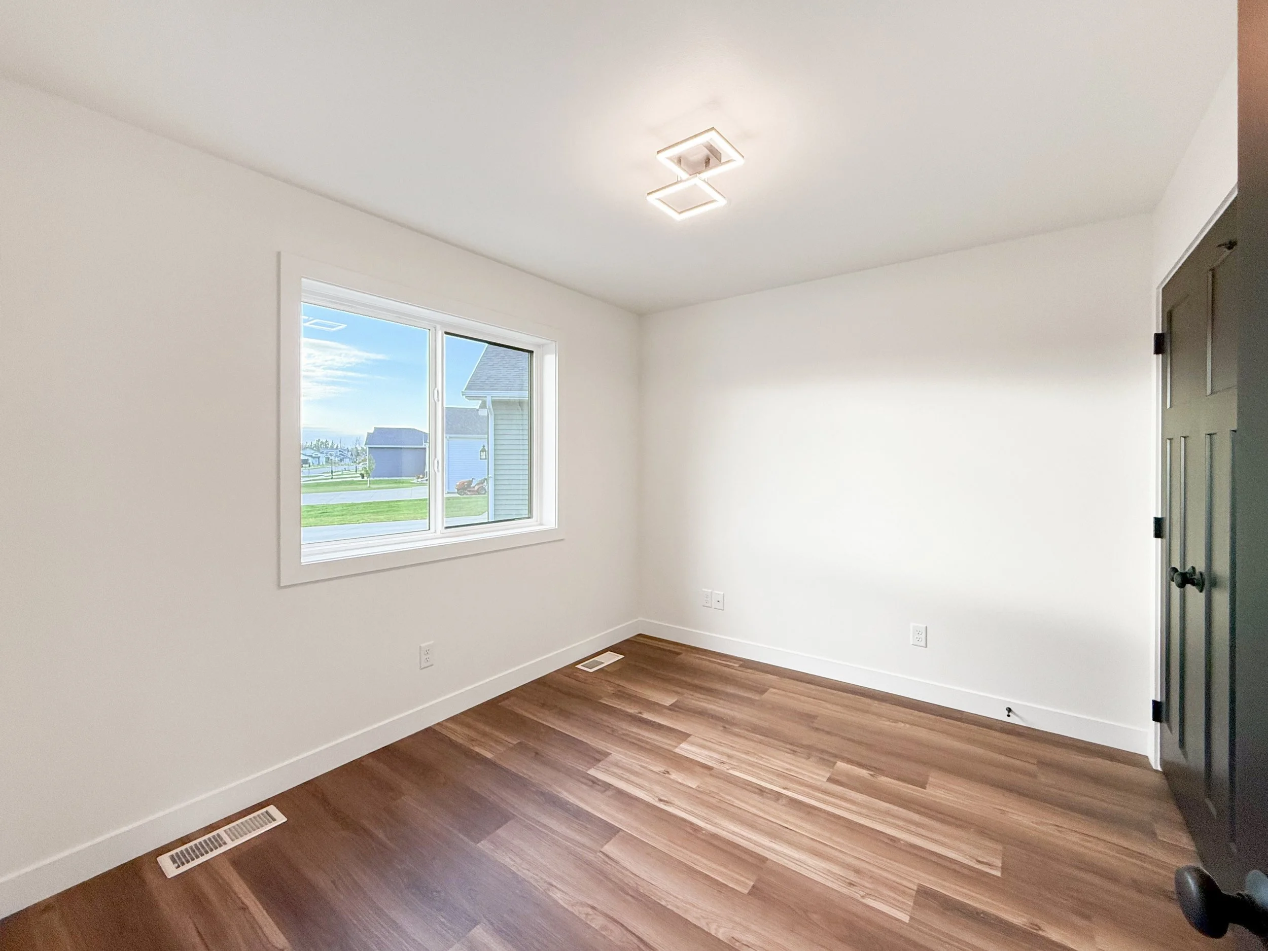 Empty room with white walls, hardwood flooring, a window showing a grassy yard and neighbor houses, black closet door, and ceiling light fixture.