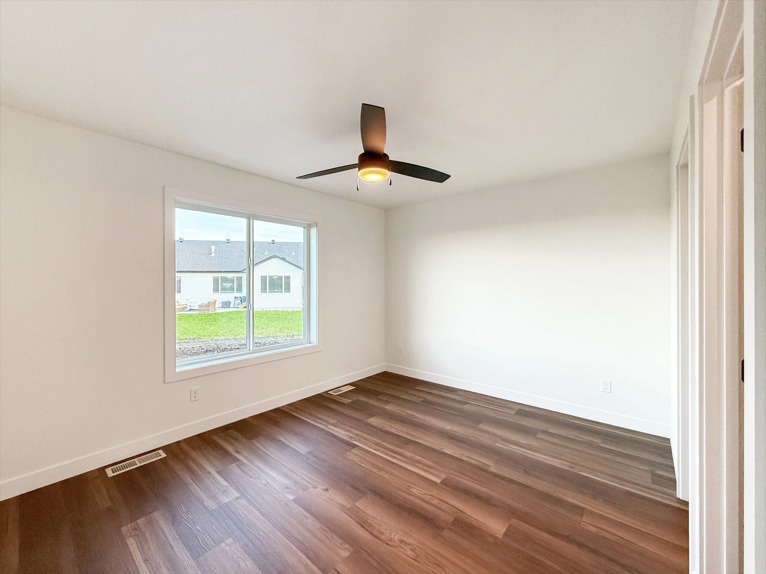 Empty room with hardwood floors, white walls, a large window showing neighboring houses, a ceiling fan with light, and a closet door on the right.