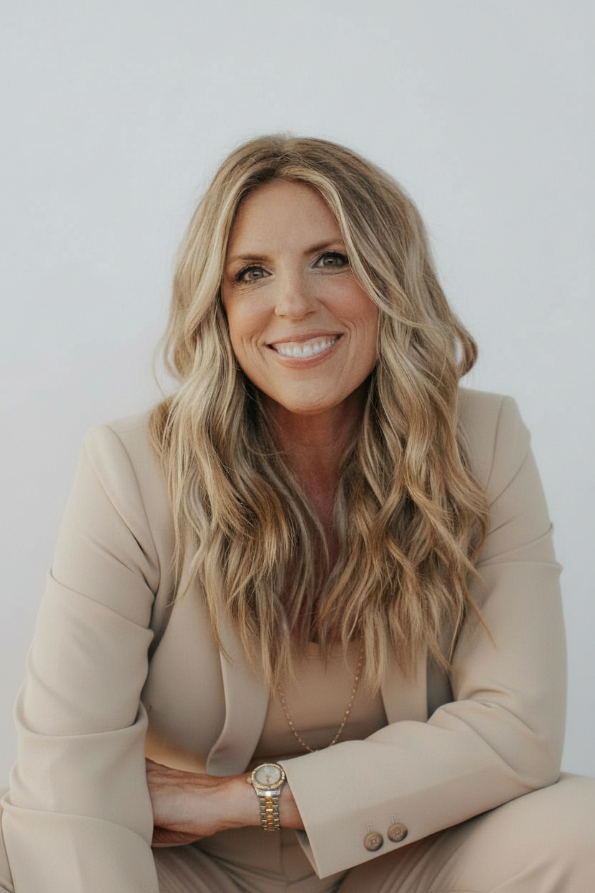 A woman with long wavy blonde hair, wearing a bright pink blazer and orange top, smiling at the camera against a plain light background.