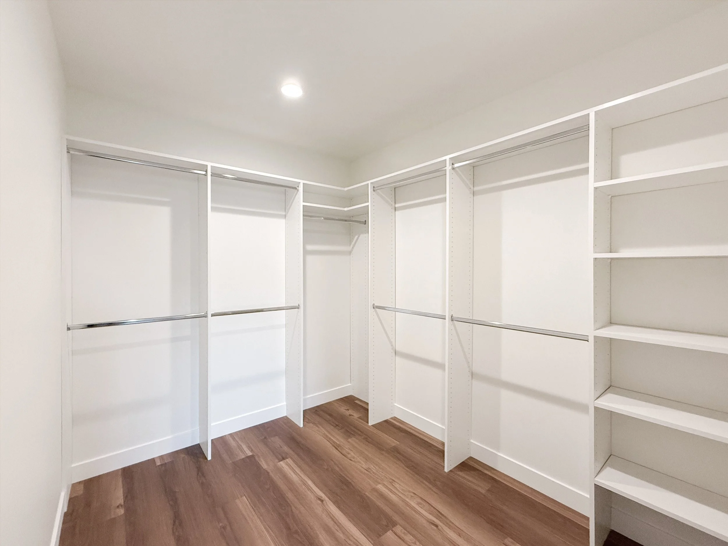 Empty walk-in closet with white shelving and hanging rods, wood flooring, and a ceiling light.