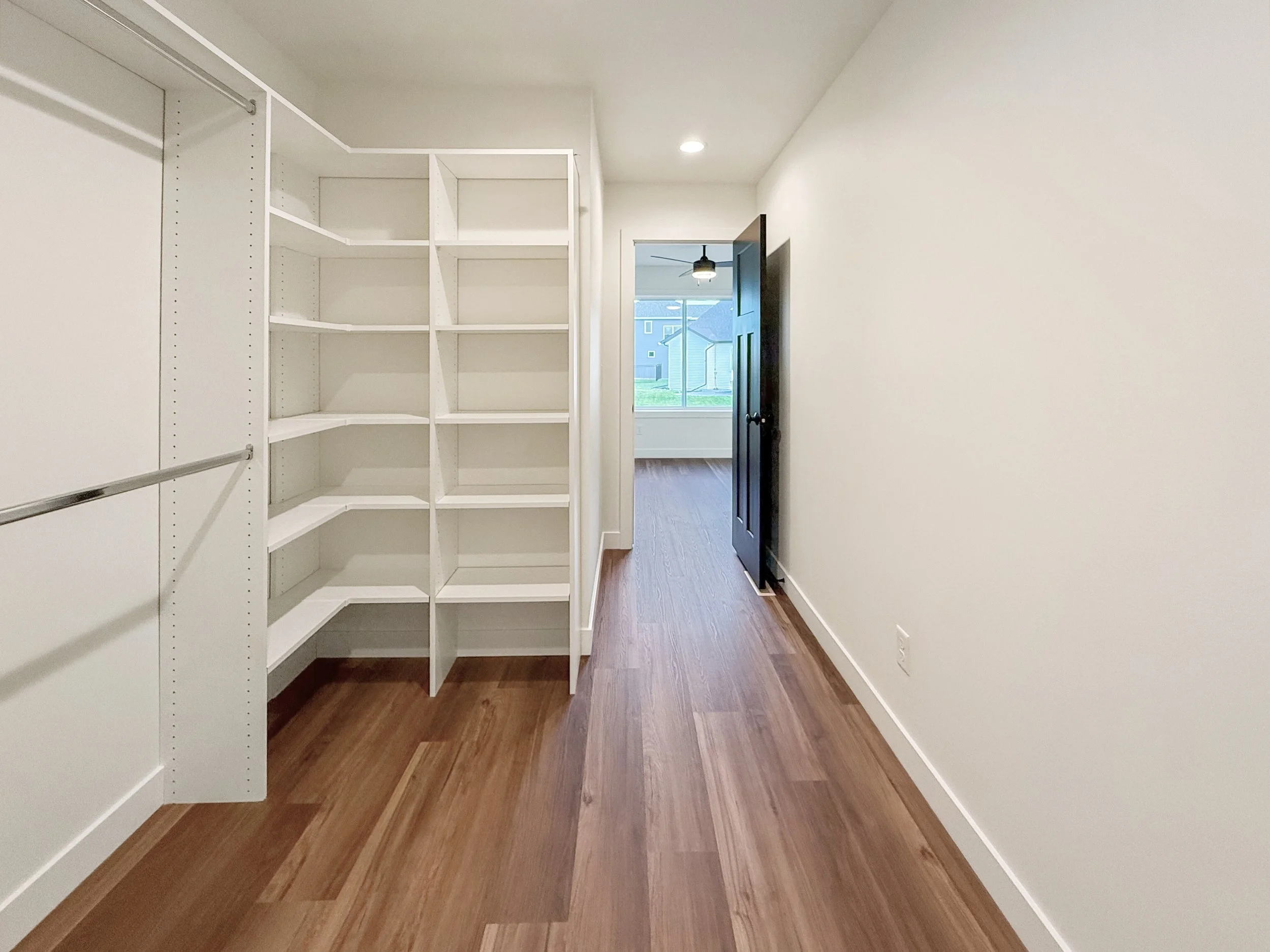 Empty walk-in closet with white shelving and wood flooring, open door leading to a room with a ceiling fan and large window.
