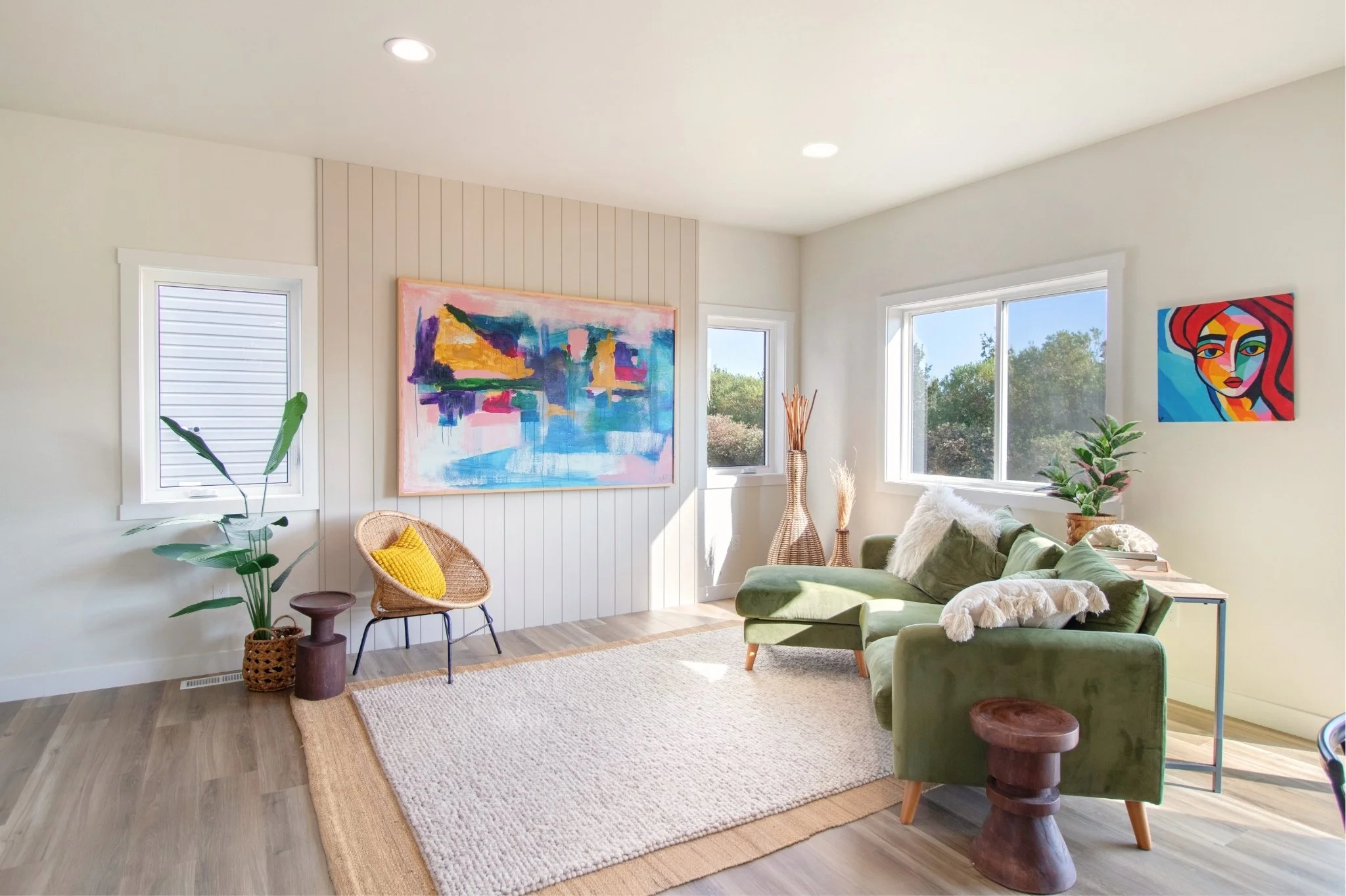 Sunlit living room with green sectional sofa, decorative pillows, wall art, potted plants, wicker chair, and wooden side table.