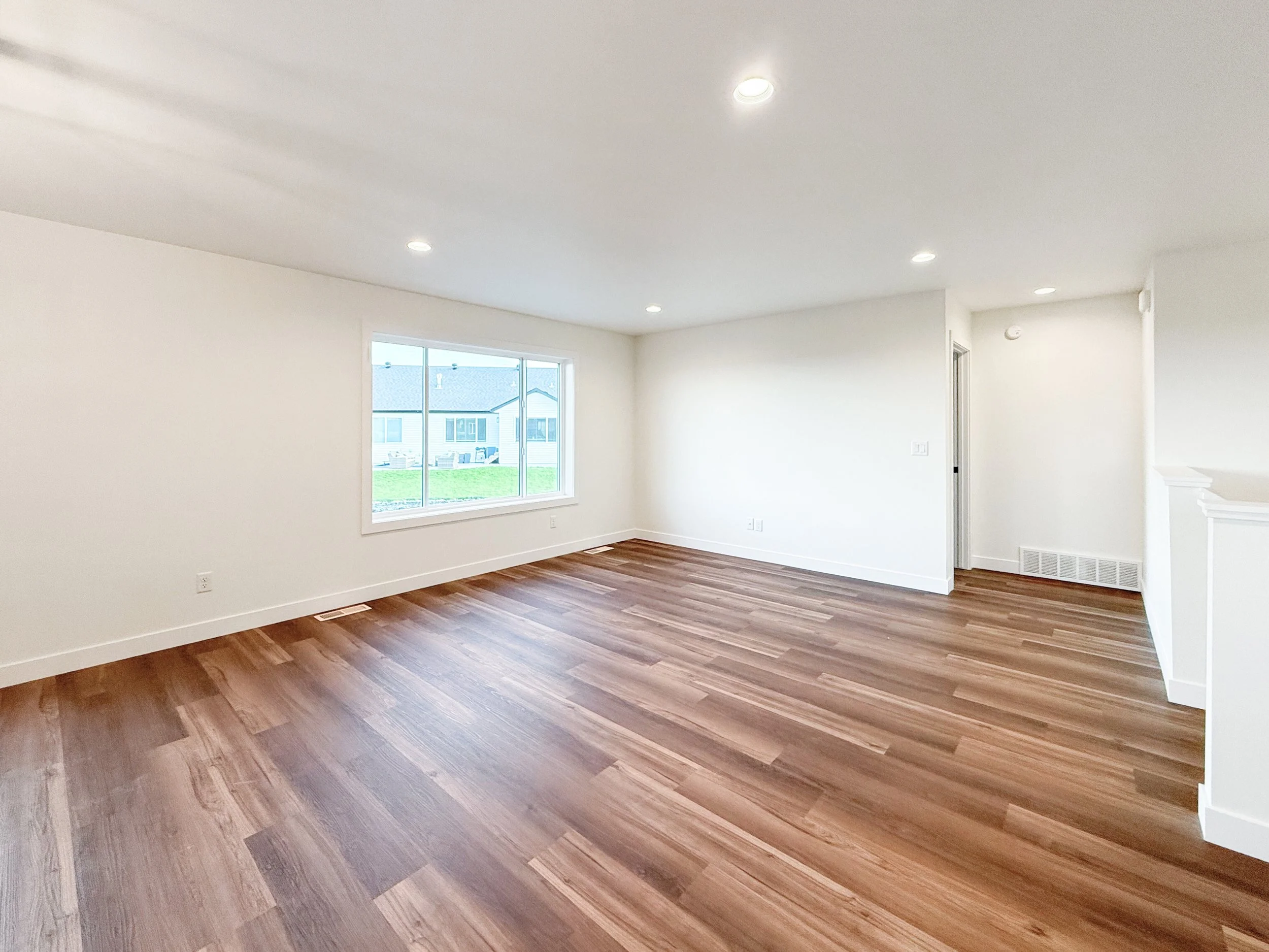 Empty living room with white walls, hardwood floor, a large window, and recessed ceiling lights.