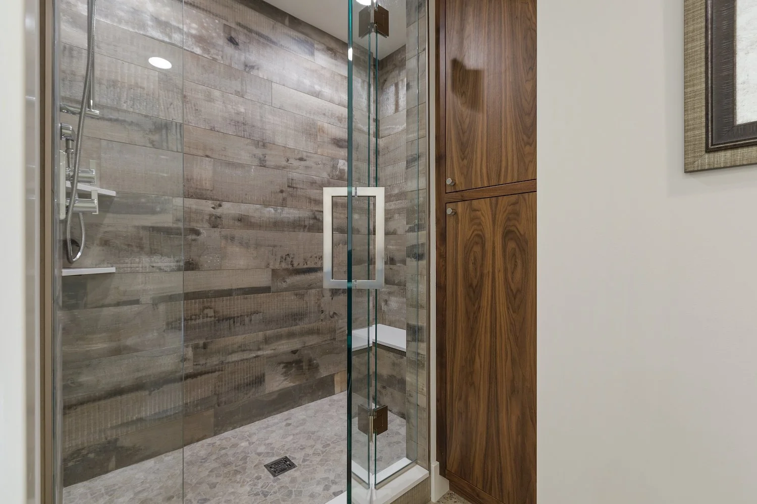 Modern bathroom shower with glass door and wood panel wall, white bench, and beige tile floor.