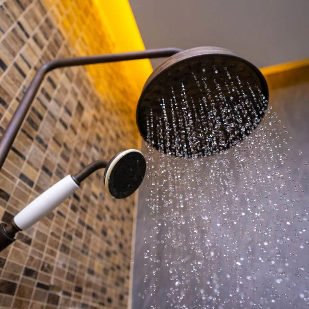 Shower with a rain shower head and a handheld shower head, water flowing from both, tiled walls in a bathroom.