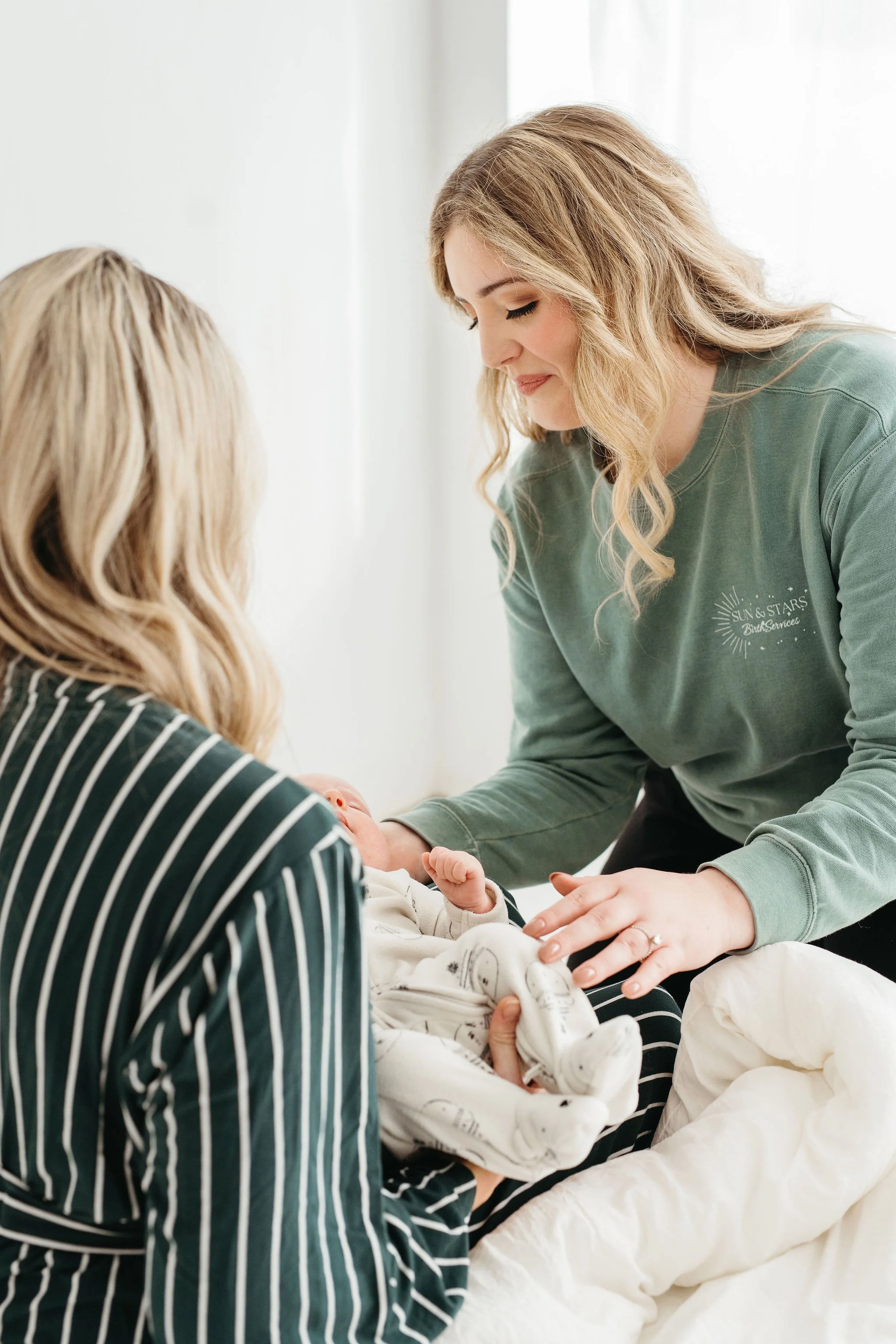 Two women holding a newborn baby in a bright room, one woman wearing a striped top and the other wearing a green sweatshirt.