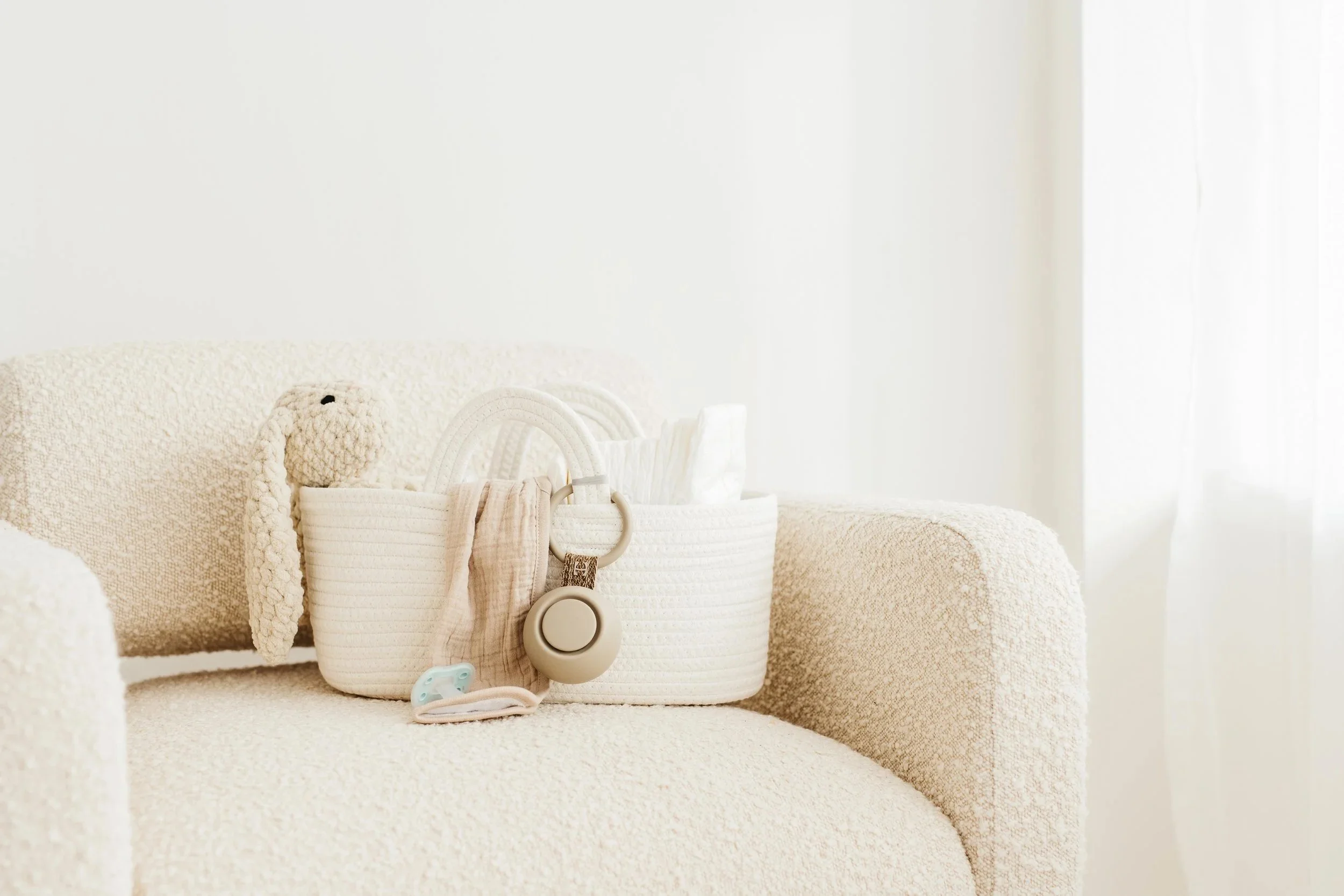 A beige armchair with a plush teddy bear and a woven basket holding baby items on it, set in a room with white walls and curtains.