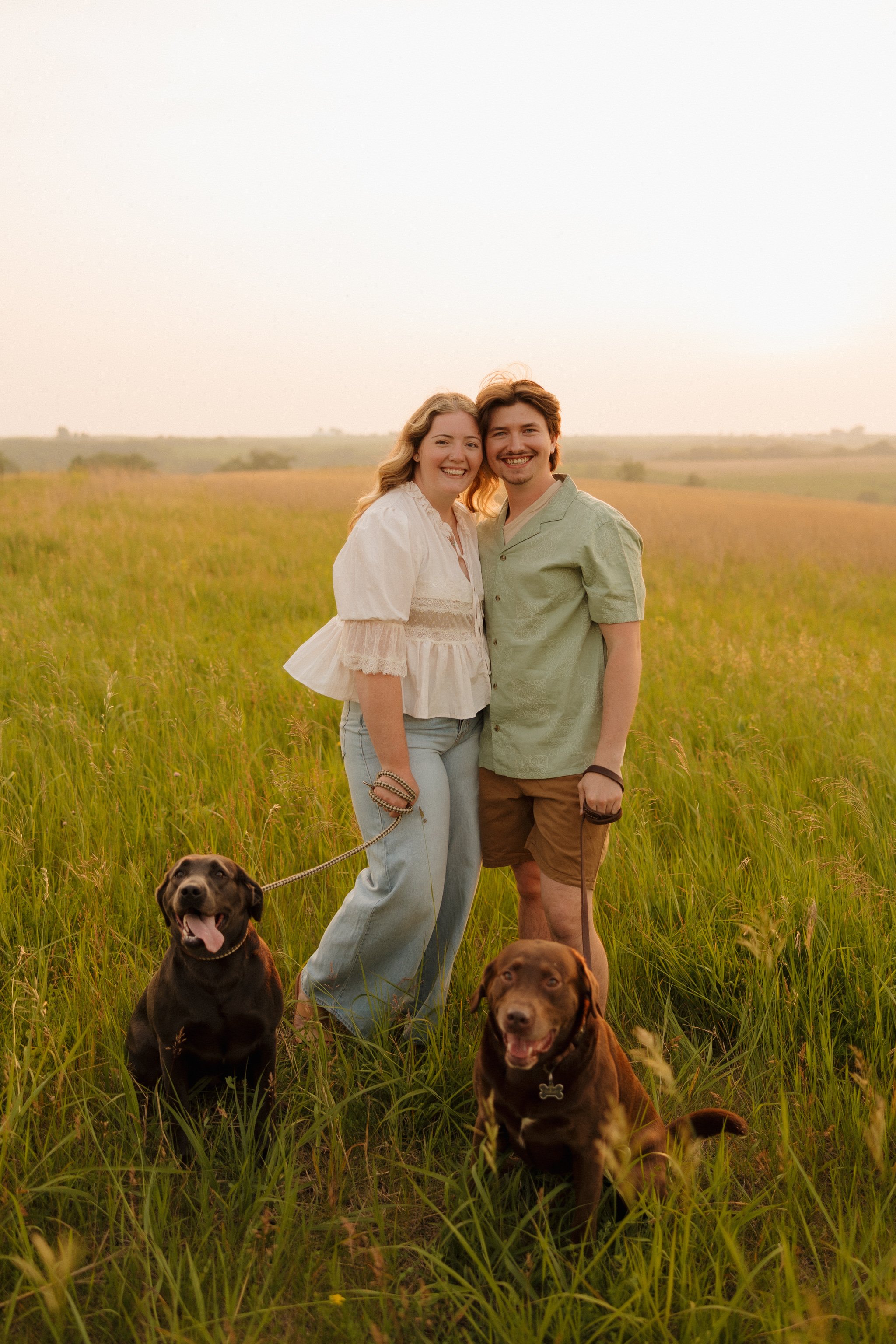 A young couple smiling and standing together in a grassy field during sunset, accompanied by three dogs, with a vast open landscape in the background.