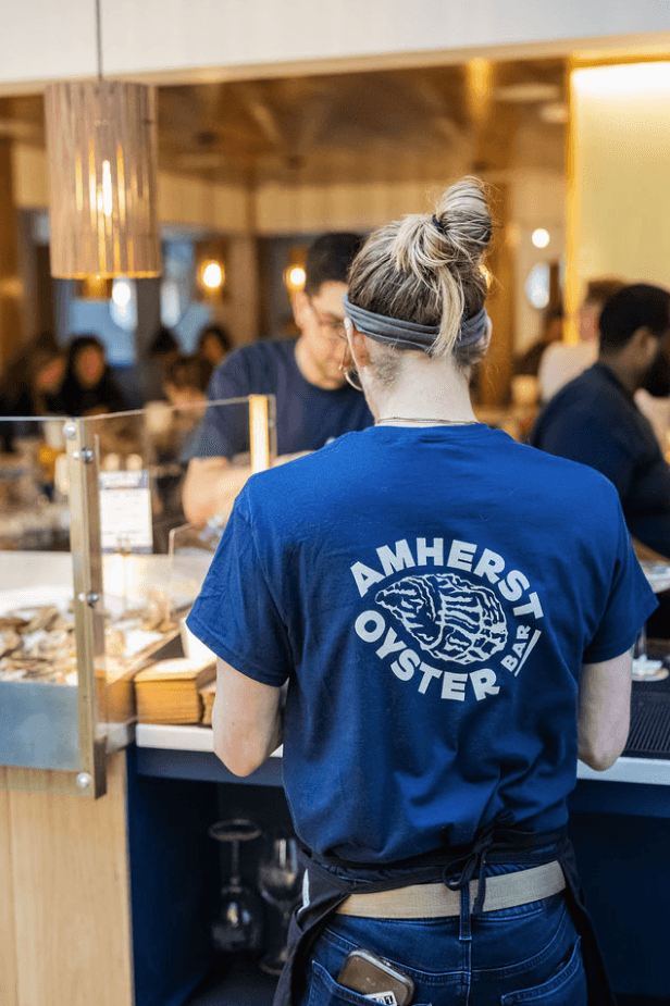 A person with blonde hair tied in a bun, wearing a headband and a blue Amherst Oyster Bar logo T-shirt, working at a busy restaurant counter with customers and staff in the background.