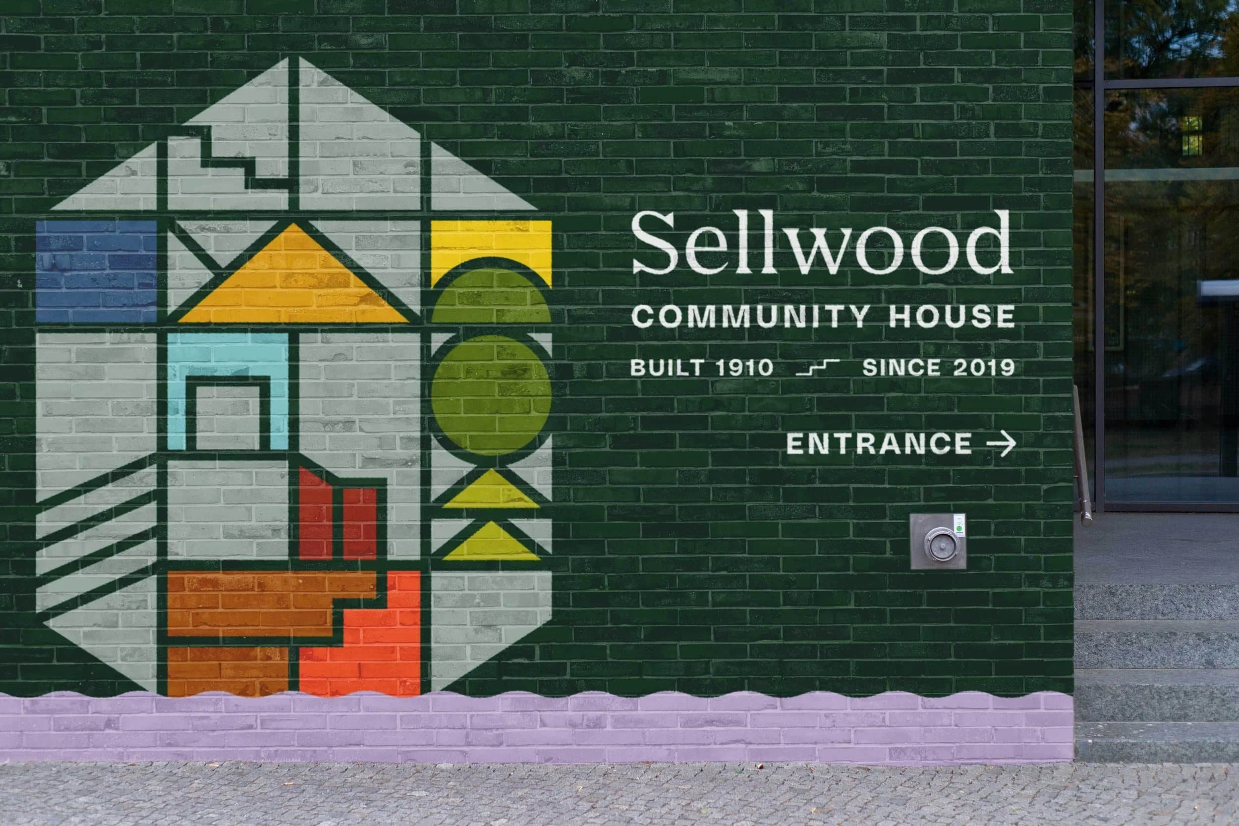 Colorful mural of a house with various geometric shapes and colors, on a dark green brick wall, with text indicating 'Sellwood Community House, Built 1910, Since 2019, Entrance'.