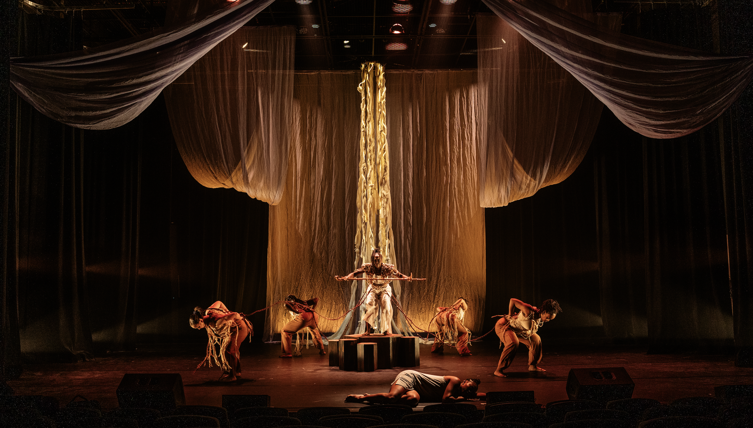 A theatrical stage performance featuring multiple dancers dressed in costume, with a backdrop of draped fabric and stage lighting.