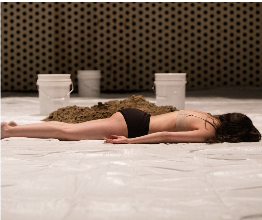 A woman lying face down on a white sheet with dirt and three white buckets in the background.