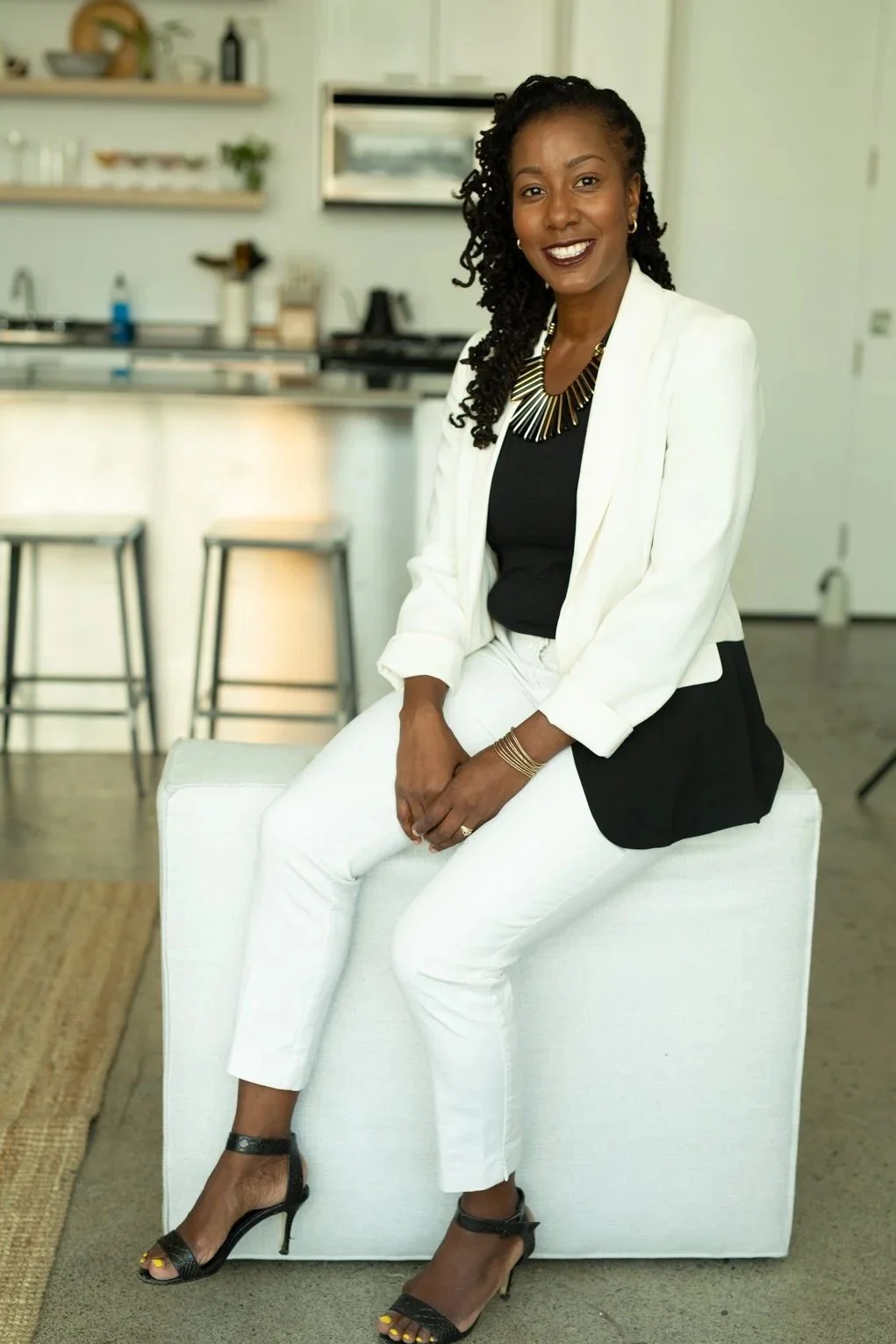 A smiling woman in a white blazer, black top, and white pants sitting on a white ottoman in a modern kitchen, wearing black high heels and gold jewelry.