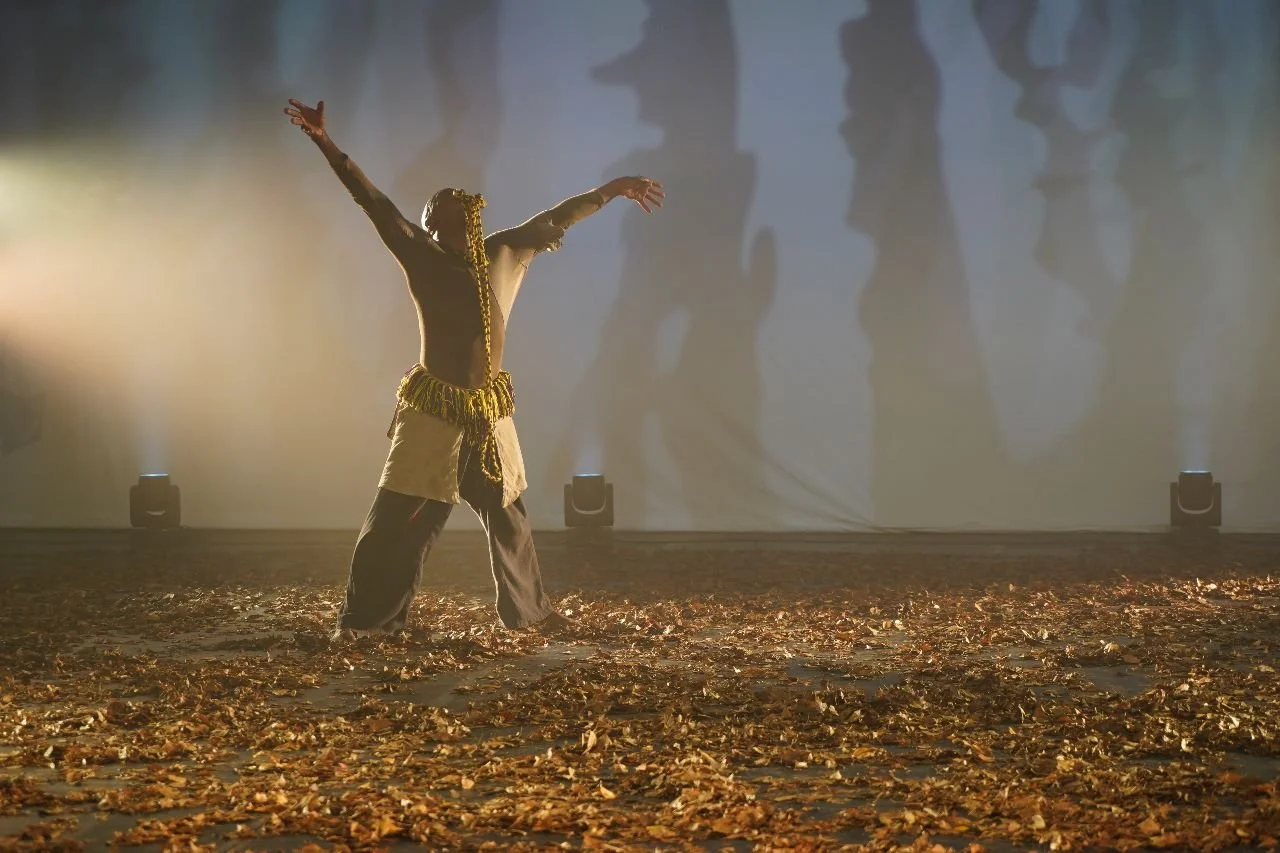 A person performing a dance or theatrical act on stage, with arms outstretched, surrounded by fallen leaves, and a foggy background with shadowy figures.