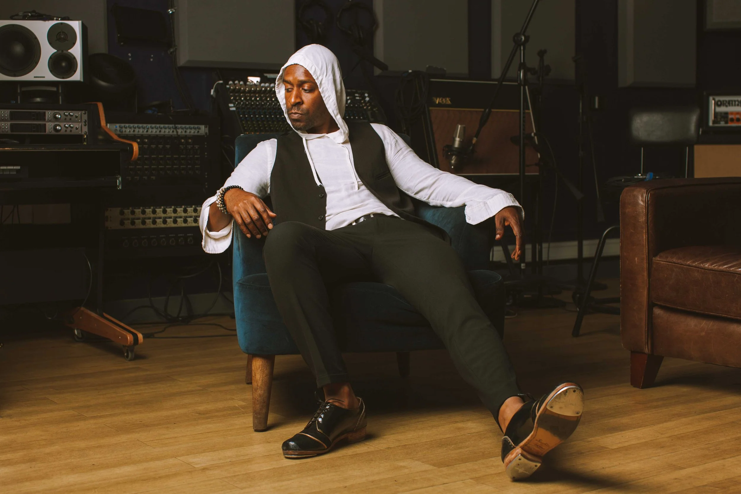 A man sitting in a chair in a music studio, wearing a white hoodie, black vest, and black pants, with a serious expression, surrounded by audio equipment and musical instruments.
