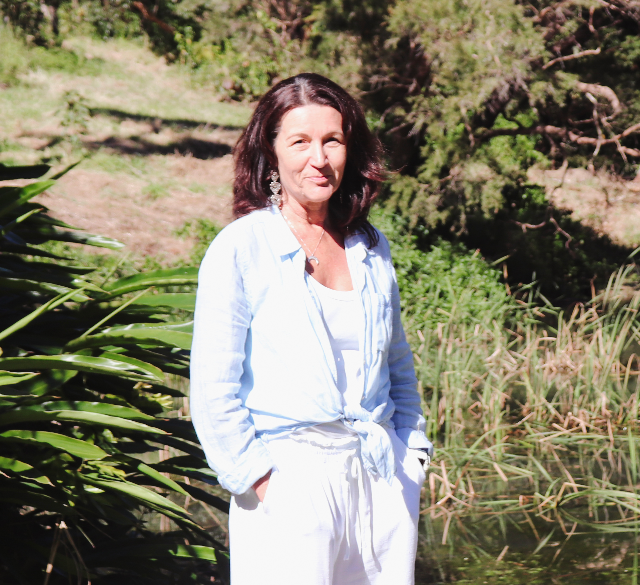 Gemma Massage Therapist with dark hair wearing a white button-up shirt and white pants stands near a pond with green foliage in the background.