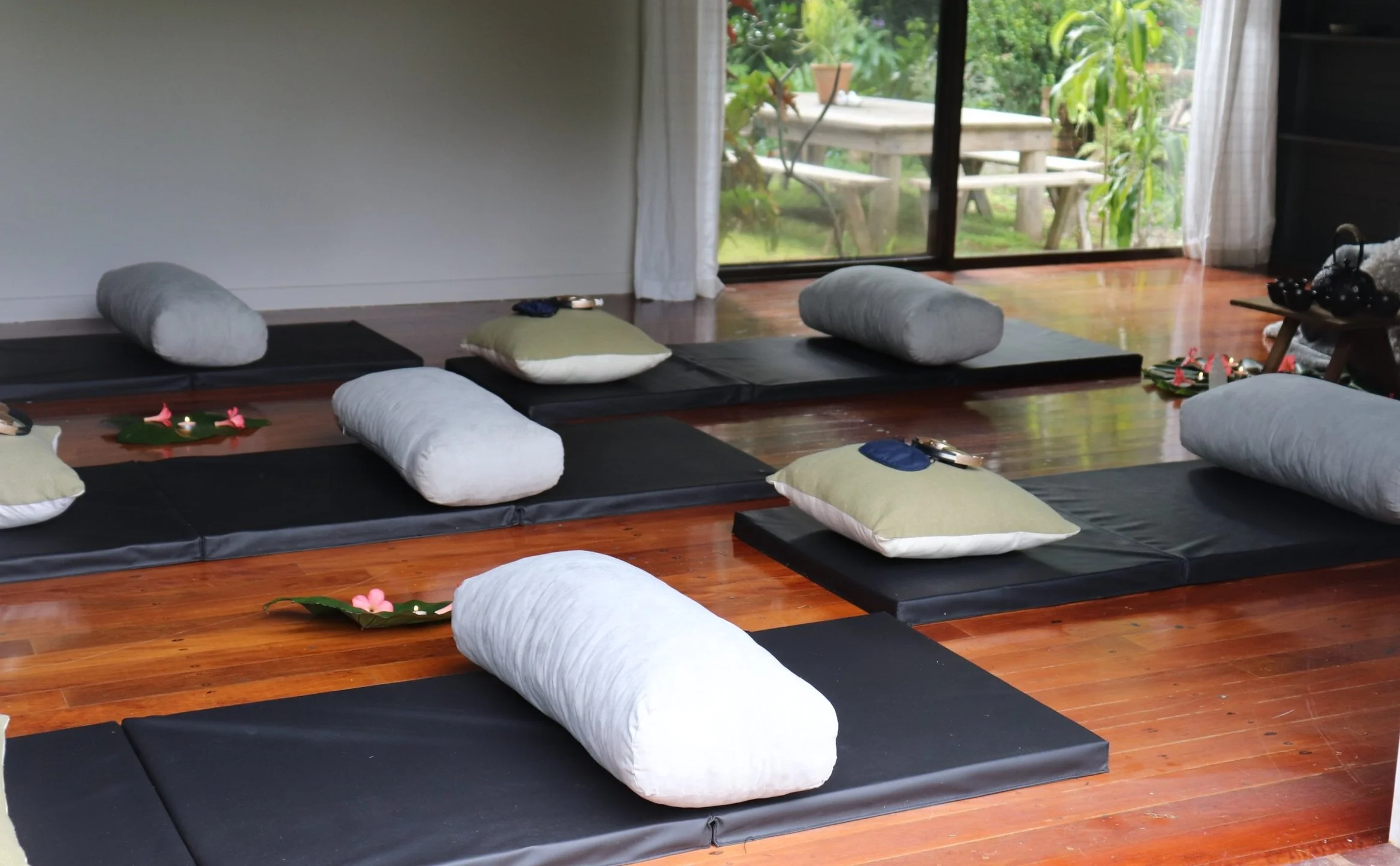 A room with meditation mats, bolsters, and cushions arranged on a wooden floor, with curtains and a sliding glass door leading outside to a garden with trees and a wooden table. Rise Temple Wellness Retreat Centre studio Byron Ba.y 