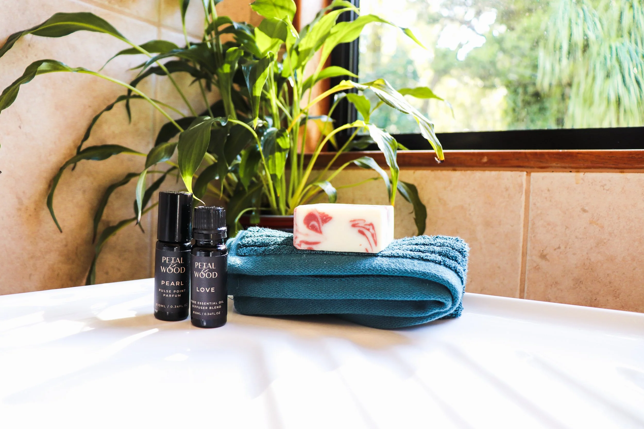 Bathroom counter with a potted plant, two Petal Wood essential oil bottles, a bar of soap with red swirls on a blue towel, and a window showing greenery outside.