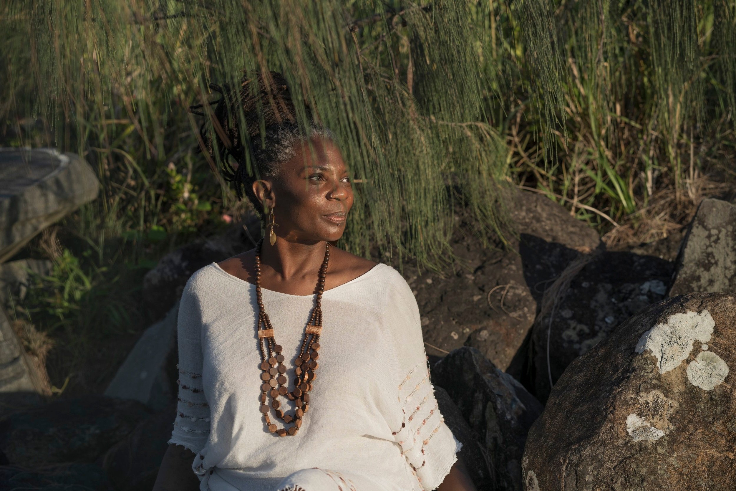 Amanda Jane Founder Rise Temple sitting outdoors on rocks near green reed plants, wearing a white dress and layered wooden bead necklaces, with her hair styled in long dreadlocks.