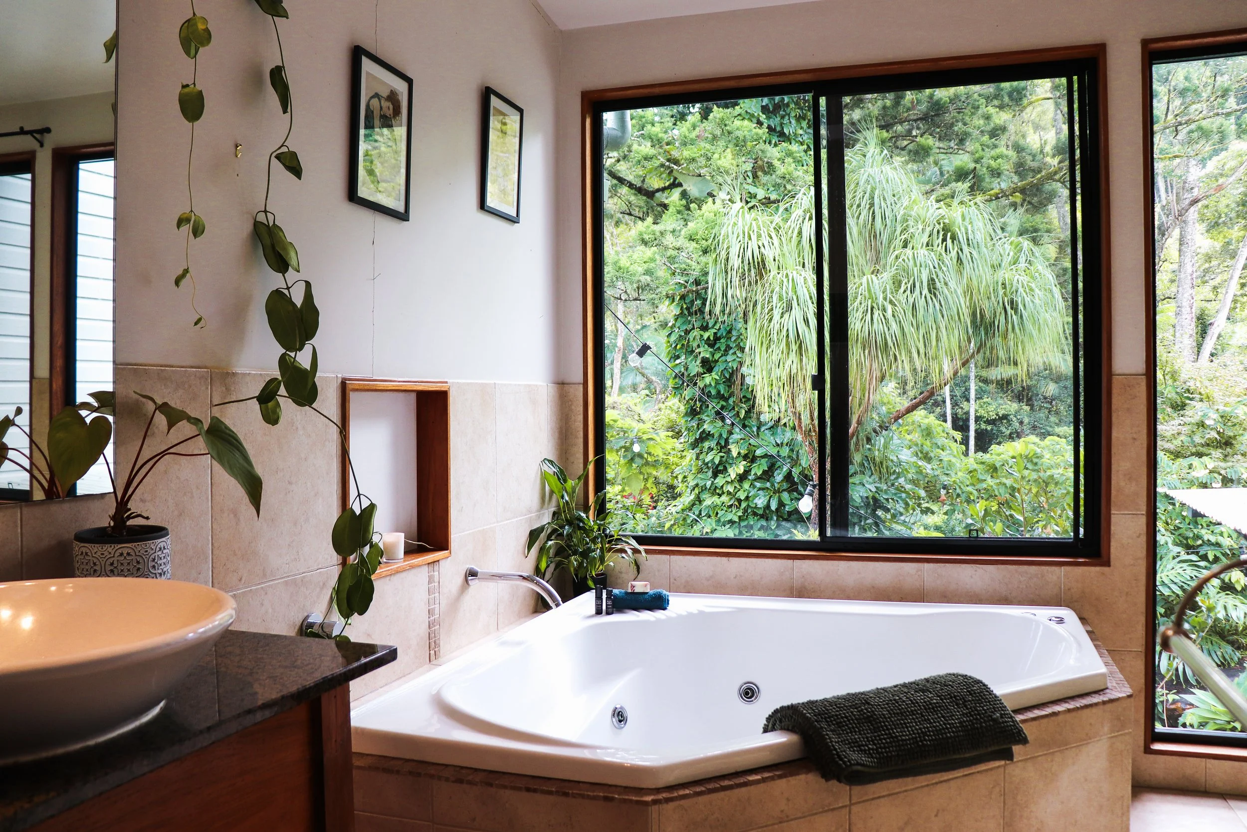 Bathroom with a large bathtub by a window showing lush green trees outside, with potted plants and framed pictures on the wall.