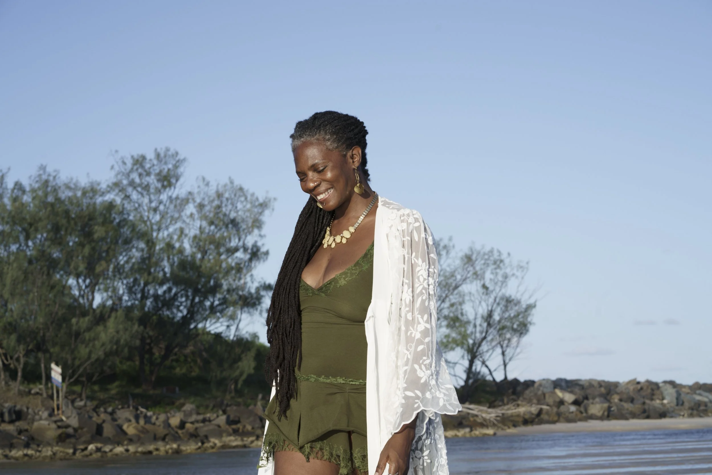 Smiling woman with long dreadlocks wearing green lace lingerie and a white embroidered cover-up, standing on a beach with rocks, trees, and a blue sky in the background.