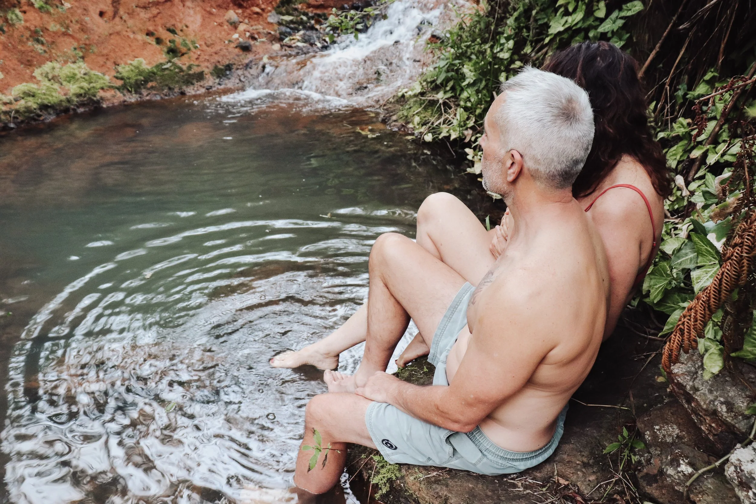 An older man with gray hair and a woman with long dark hair sitting on a rock at the edge of a small, flowing creek, surrounded by green foliage at Rise Temple Retreat.