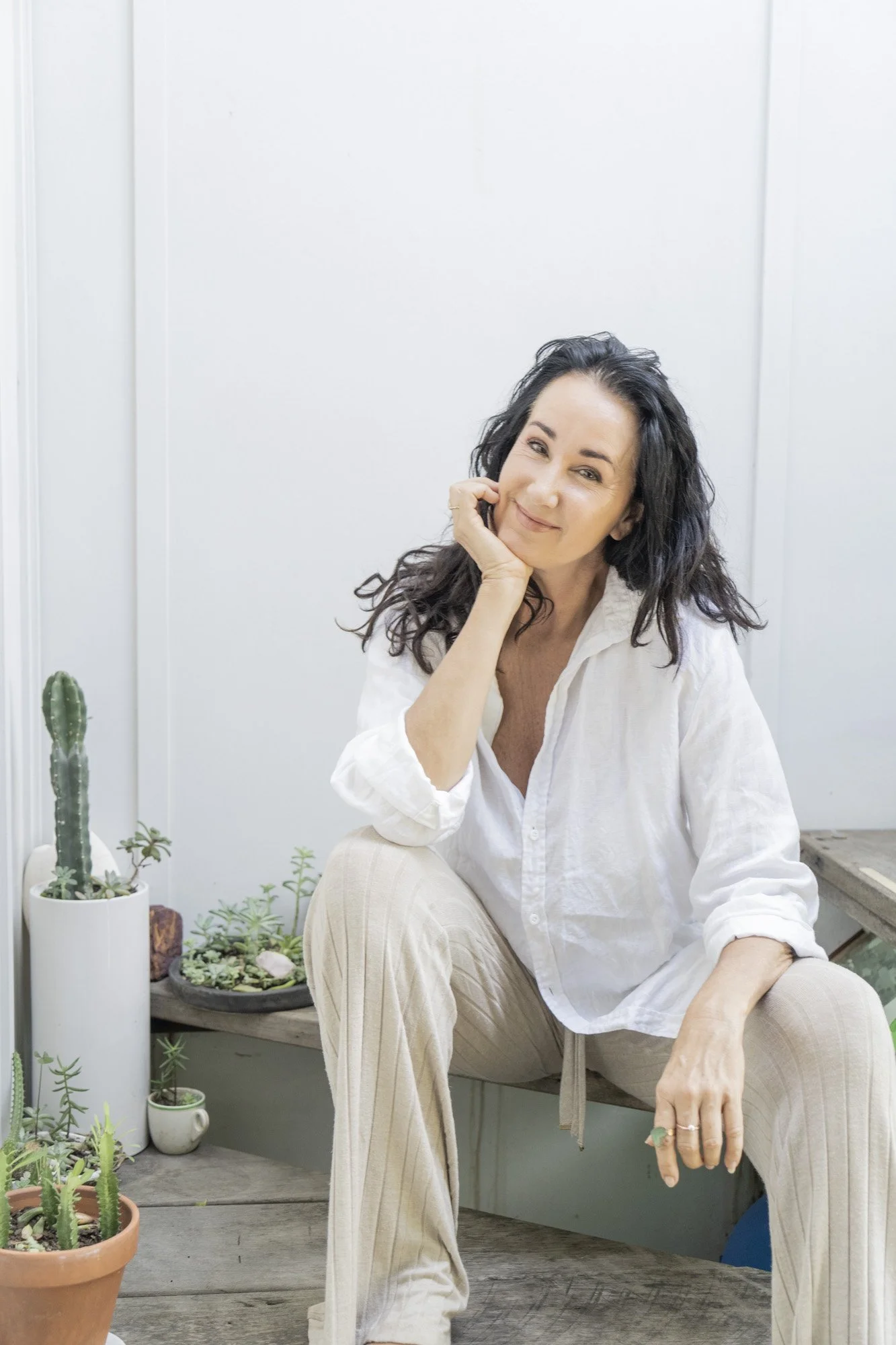 Deon Demouche Counsellor, curly hair, wearing a white shirt and beige pants, sitting on a wooden surface with small potted succulents and cacti around her, smiling with one hand resting on her cheek, in a bright room with white walls.