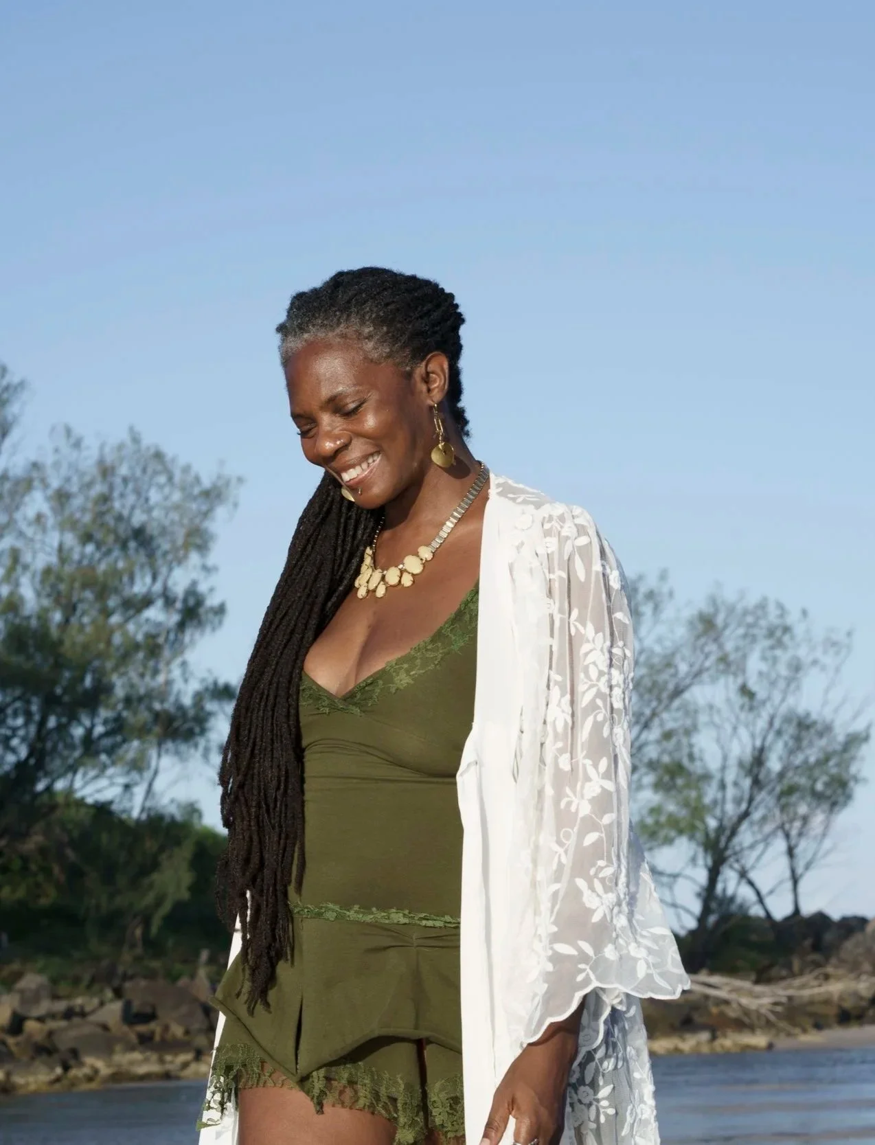 Amanda Jane founder of Rise Temple smiles downwards while standing outdoors near a body of water with trees in the background. She is wearing a green lace camisole, matching shorts, a white sheer embroidered cover-up, and gold jewelry.