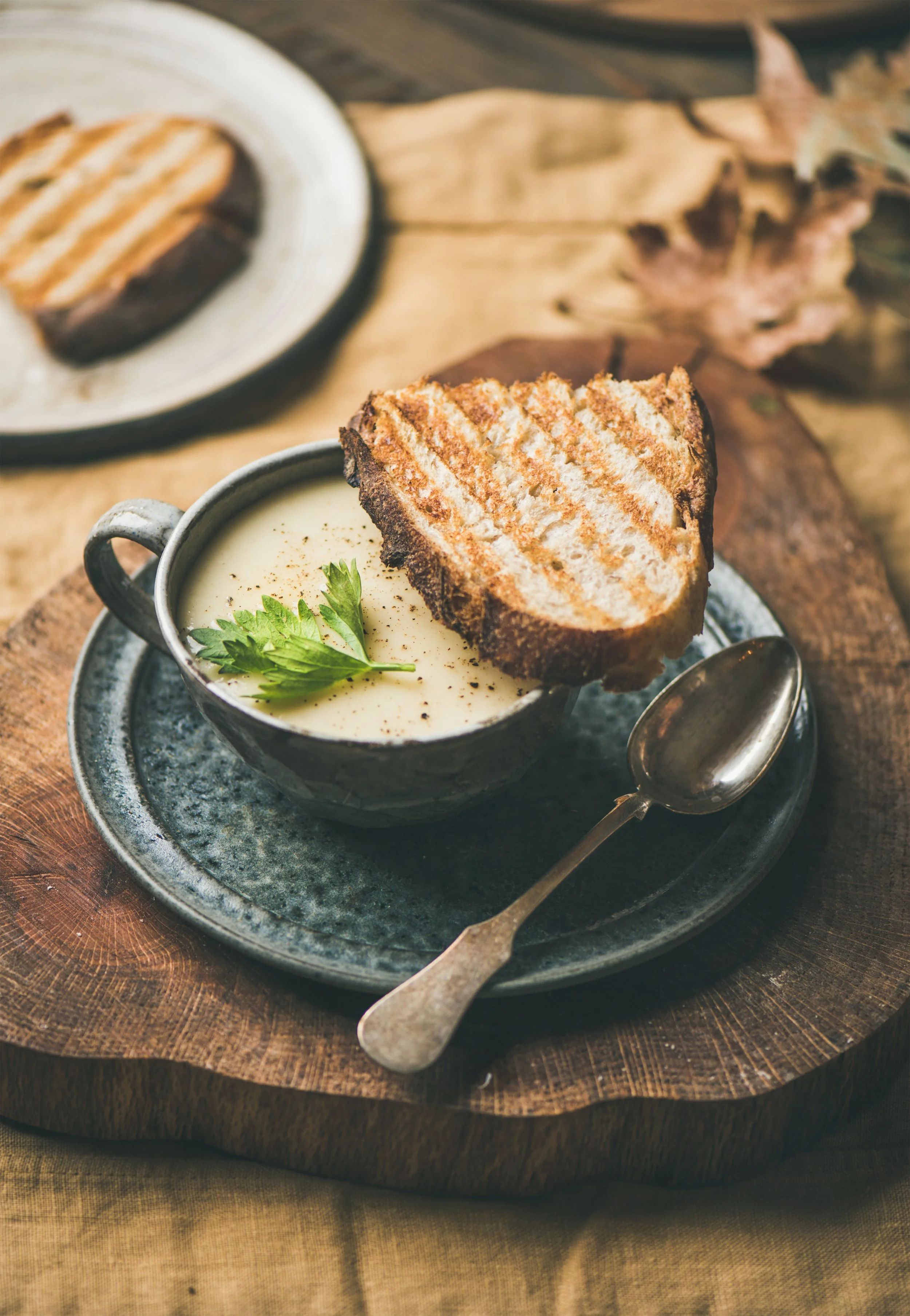 A bowl of creamy soup garnished with a green parsley leaf and black pepper, served with a grilled cheese sandwich on a rustic wooden tray.