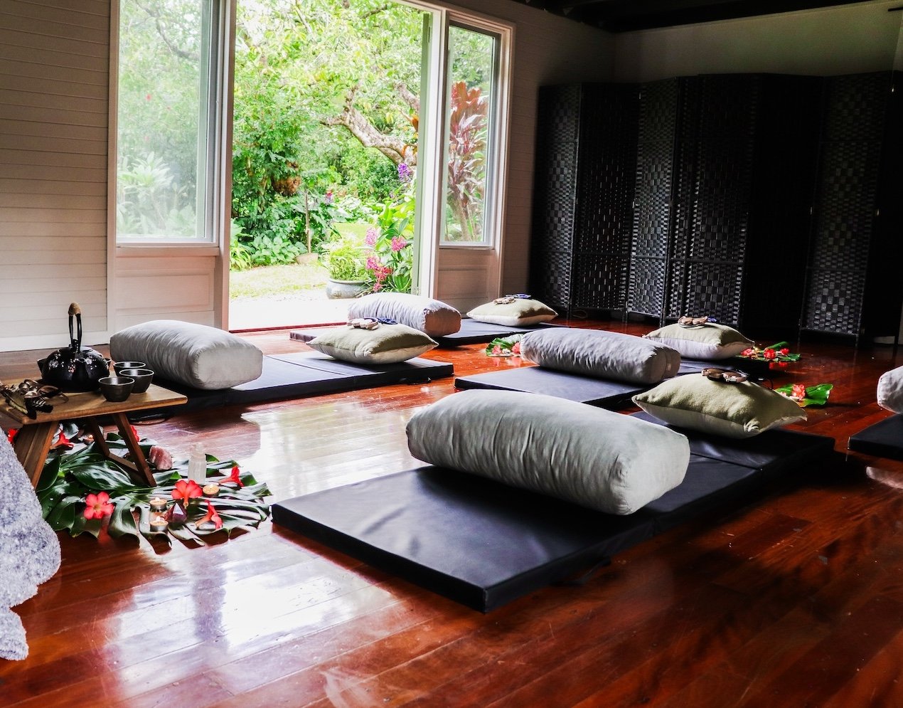 Room set up for meditation or yoga with black mats, white bolsters, and pillows, open to a view of a lush garden with plants and trees. Rise Temple Wellness Retreat Centre Byron Bay Studio.