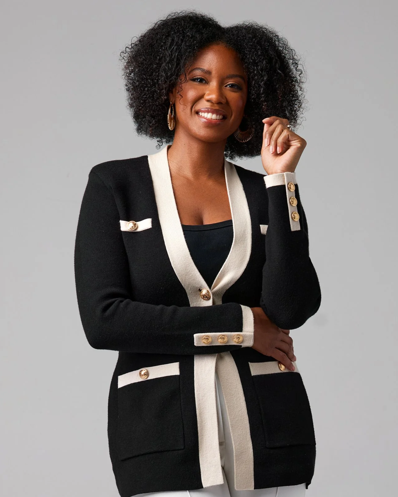 A Black woman with curly hair smiling, wearing a black and white blazer with gold buttons and gold earrings, standing against a plain gray background.
