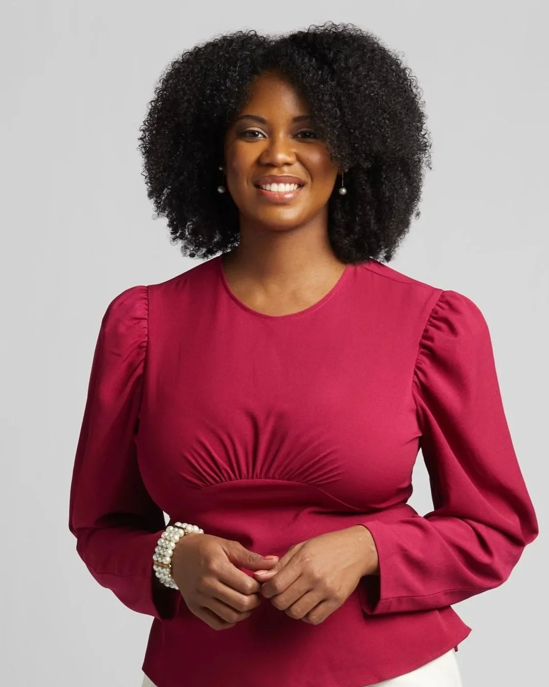A woman with curly hair wearing a red blouse with puffed sleeves and pearl earrings, smiling against a light gray background.