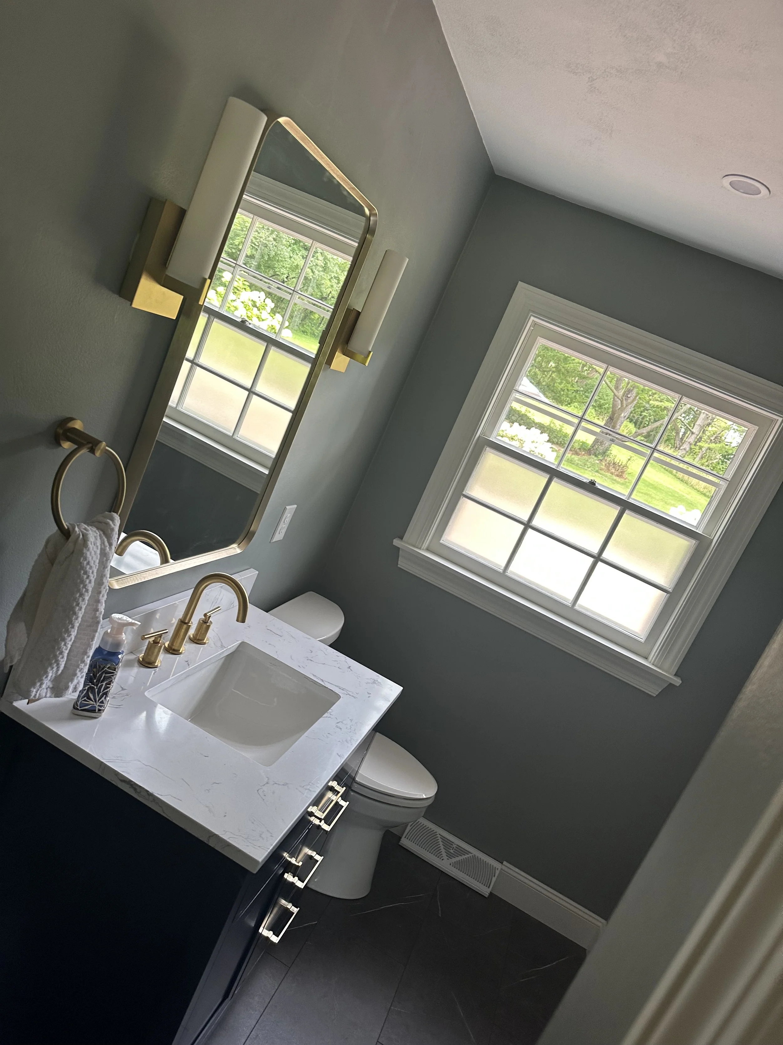 A modern bathroom with a white marble sink countertop, brass fixtures, a large rectangular mirror, and two tall wall-mounted lights. There's a window showing a green outdoor scene, and a white toilet below the window. The flooring is dark tile, and t