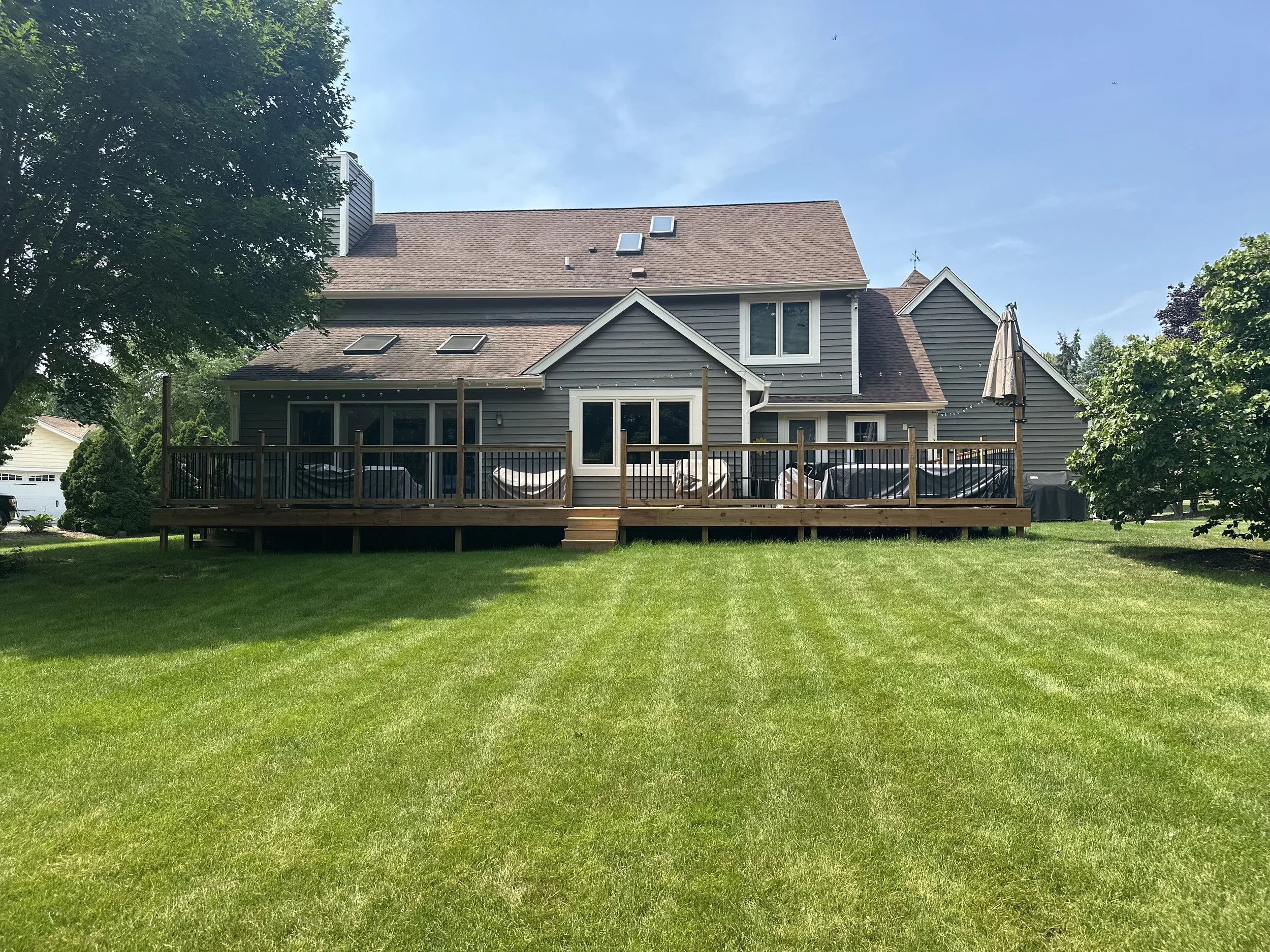 Backyard with a wooden deck, surrounding green lawn, trees, and a house with gray siding and a brown roof under a clear blue sky.