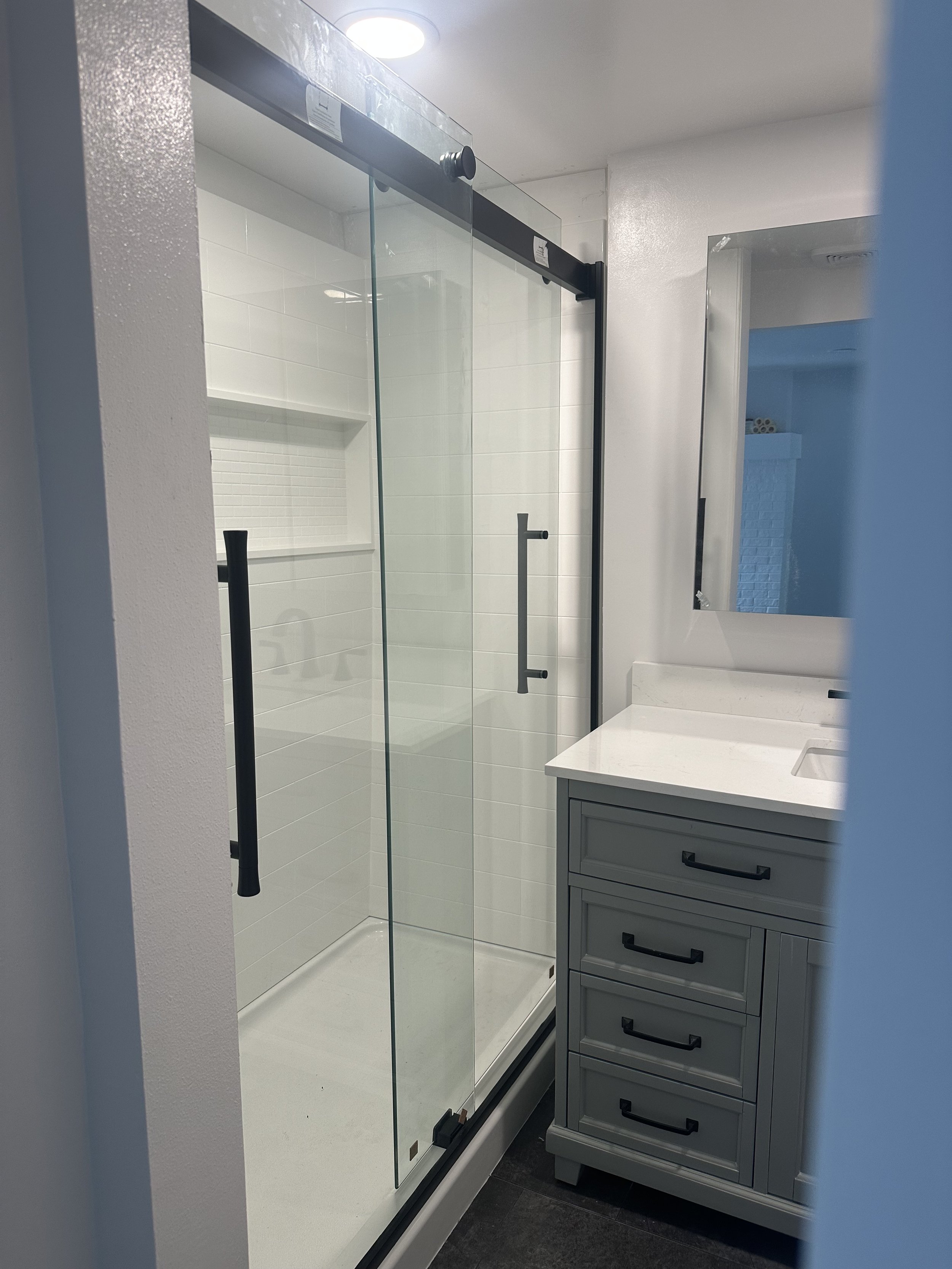 A bathroom with a glass shower enclosure, a vanity with a white countertop, and a grey cabinet with black handles.