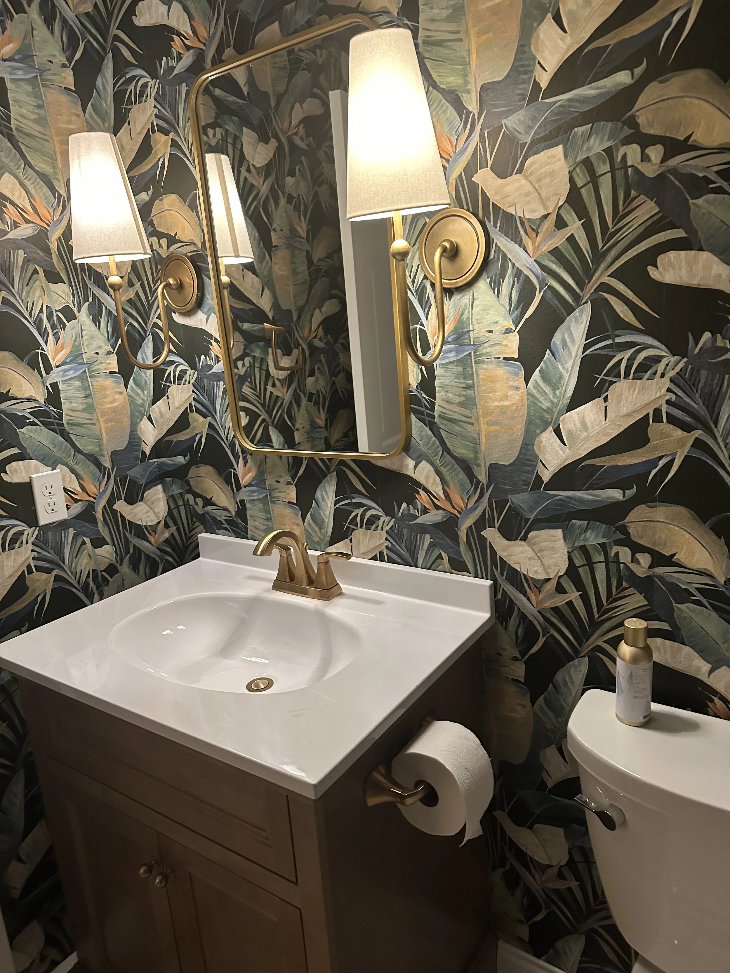 A bathroom vanity with a white sink, brass fixtures, a framed mirror, and a tropical leaf wallpaper. There is a bottle on the side table and a toilet paper roll underneath the sink.