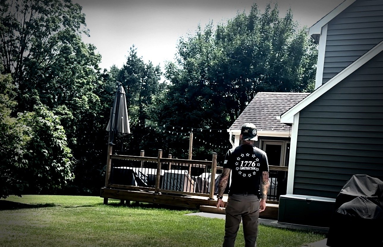 A man in a black t-shirt and baseball cap standing in a backyard near a house, with a deck and patio furniture visible. Trees and sky are in the background.