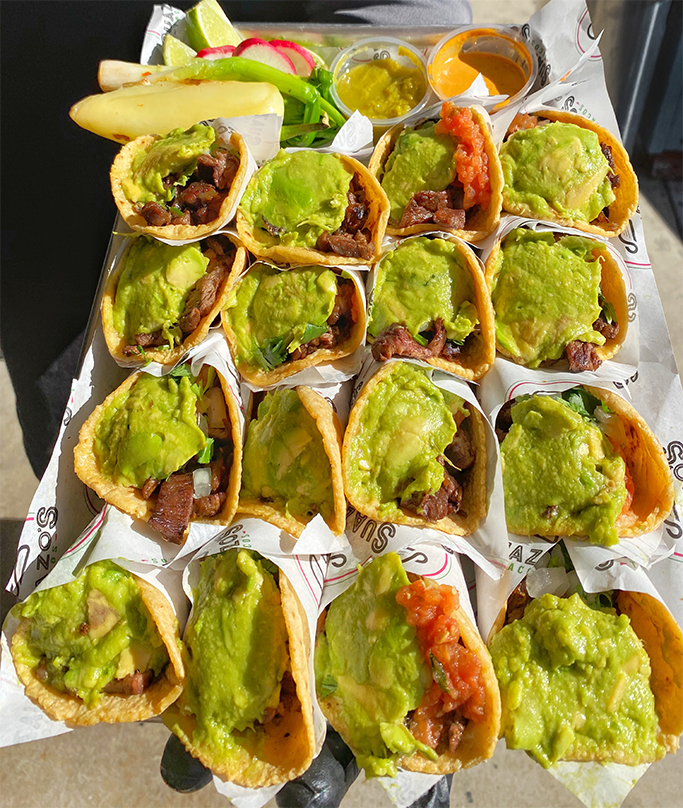 Tray of tacos filled with beef and topped with guacamole, served with lime wedges, radishes, and a banana, with salsa and hot sauce in small containers.